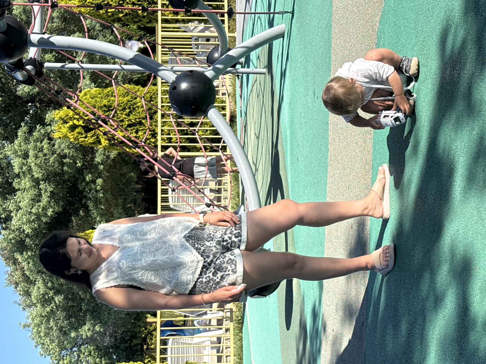 A woman stands in a playground, looking down at a child. The woman is wearing a white sleeveless top with intricate embroidery and leopard print shorts. She has dark hair and is wearing tan sandals. The child, wearing a white hooded shirt and shorts, is crouched on the ground, playing with a toy ambulance. The playground features a large, grey and black climbing structure with pink ropes. The ground is covered in a green and light-colored rubberized surface. In the background, a yellow fence surrounds lounge chairs and trees. The scene appears to be an outdoor recreational area, possibly in Sochi, Russia, given the sunny weather and lush greenery, consistent with coastal resort towns.