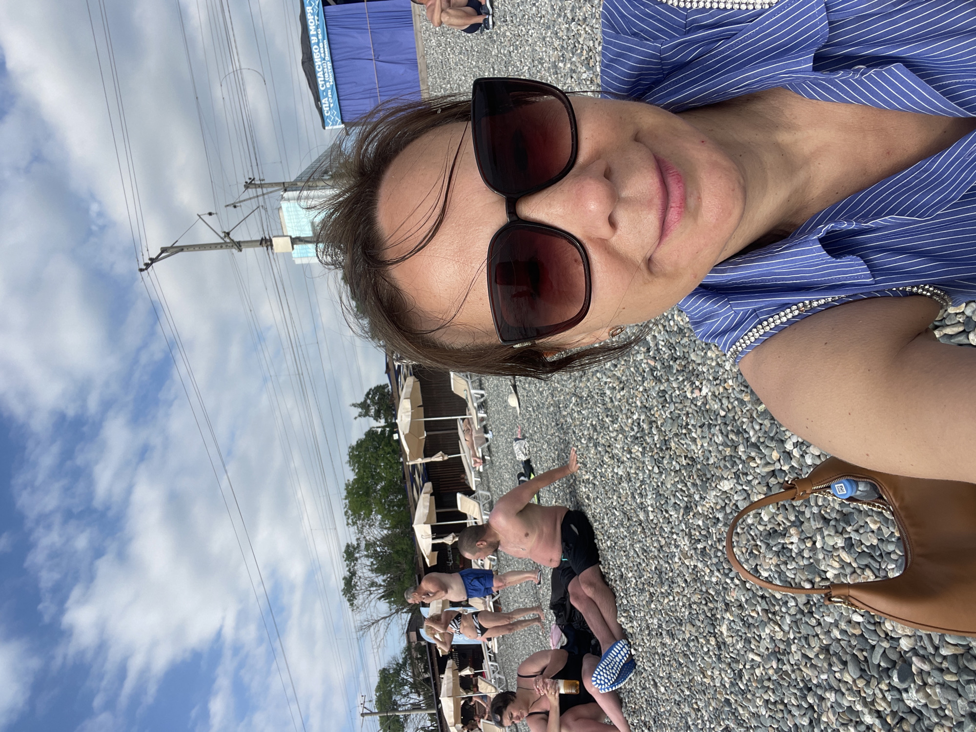 This media file captures a group of people on a pebble beach in Khostinskiy, Russia. The perspective is a selfie taken by a woman wearing sunglasses and a blue and white striped top adorned with rhinestones. She is smiling at the camera, suggesting a pleasant mood.
In the background, several other individuals are visible, some lounging on beach chairs under umbrellas, while others are standing or sitting on the pebbled shore. One man in black shorts is in the foreground, appearing to be stretching or exercising. The scene suggests a relaxed, leisurely day at the beach.
The weather appears to be cloudy with patches of blue sky. Overhead, power lines and pylons are visible, indicating proximity to a railway or an electrical infrastructure. Behind the beachgoers, there are some buildings or structures, one of which has a blue awning with visible text. The text reads "СПА - СПАСИБО У МОРЯ" which translates to "SPA - THANK YOU BY THE SEA," along with a phone number and website.
The time of day is likely afternoon, given the lighting conditions and the activities of the people. The overall ambiance is one of a typical summer day at a Russian seaside resort. A brown handbag with a blue water bottle peeking out of a zippered compartment rests on the pebbles near the selfie-taker.
