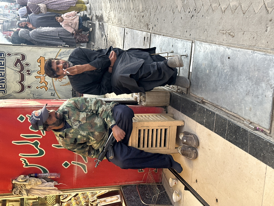 The image depicts a street scene in a commercial area of Quetta City Tehsil, Pakistan.
In the foreground, two adult males are seated side-by-side. The man on the left, with a beard and mustache, wears a camouflage jacket, dark trousers, and a dark blue "SECURITY" baseball cap featuring the Pakistani flag. He is seated on a light-colored wooden slatted stool, holding a compact black assault rifle (likely an MP5 variant) horizontally across his lap with his right hand. His brown slip-on shoes are on the ground in front of his stool, and he wears dark socks. He appears to be smiling and looking towards the right. To his left, a red shopfront wall with white Arabic script (Urdu) is visible, along with a gold jewelry display and a clear plastic bottle hanging from a hook. On the ground near him, three small teacups and a metal dish rack are present.
The man on the right, also with a beard and mustache, wears a dark, possibly black, shalwar kameez. He is seated on a simple metal-framed stool with his legs crossed, holding a lit cigarette to his mouth with his right hand, in the act of smoking. He looks generally forward or slightly towards the left. A light-colored plastic jerrycan rests on the ground beside his stool.
Both men are positioned along a demarcation line on the ground, separating an interior paved area from an exterior concrete sidewalk. In the midground, beyond the man on the right, several individuals, primarily women in traditional head coverings and outer garments, and a small child in a pink coat, are walking on the pavement. Behind them, another shopfront is visible, featuring "JEWELLERS" in both English and Arabic script (Urdu). The lighting suggests an overcast sky or diffused light conditions.