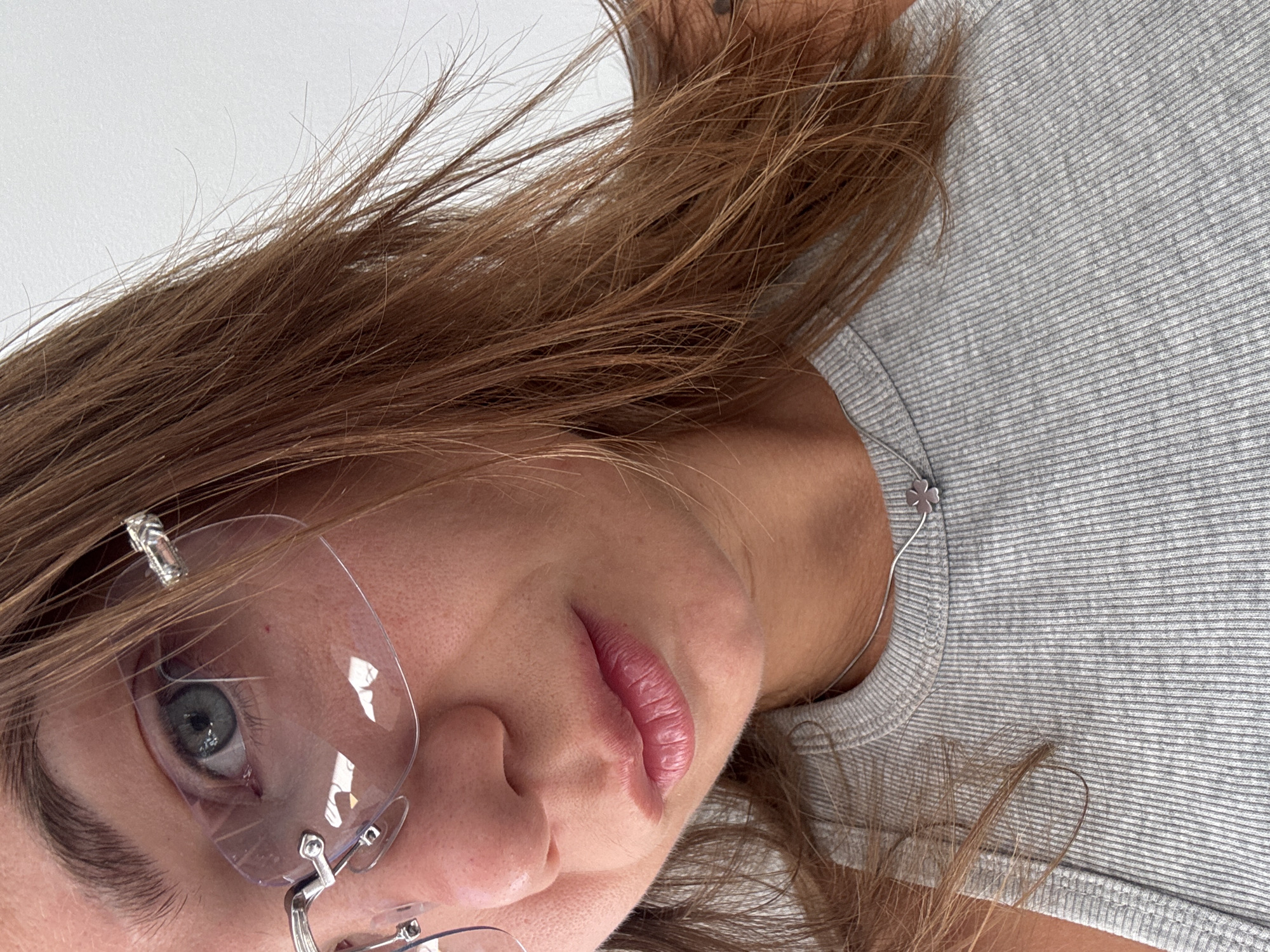 The image shows a close-up of a young woman's face and upper torso. She has light skin, hazel eyes, and long, reddish-brown hair. She is wearing clear, rimless glasses and a grey ribbed tank top. A delicate silver necklace with a clover pendant is visible around her neck. The background is a plain, white wall. There is no identifiable information that places this image specifically in Budva, Montenegro; it is a generic indoor or studio setting.