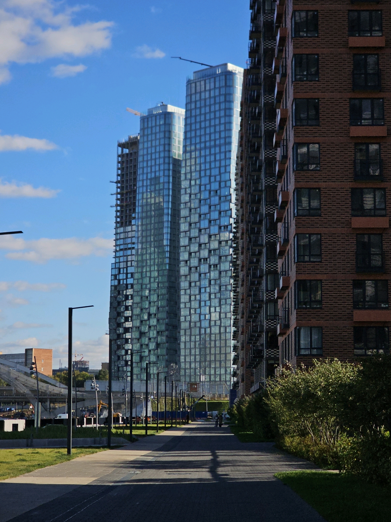 This is a photograph of the Moscow City business district in Moscow, Russia, under a bright blue sky with scattered clouds. In the foreground, a paved pedestrian walkway runs alongside a manicured green lawn. To the right of the walkway is a tall, brick-clad residential building with numerous windows and balconies, its facade casting a shadow onto the path. To the left of the walkway, a row of modern, tall glass skyscrapers dominates the skyline. One of these skyscrapers appears to be under construction, with its upper floors unfinished. In the middle ground, there are construction sites with cranes, scaffolding, and various construction equipment, suggesting ongoing development in the area. Lampposts line the walkway, and in the distance, other buildings and infrastructure are visible. Two indistinct figures are walking away from the camera on the walkway.