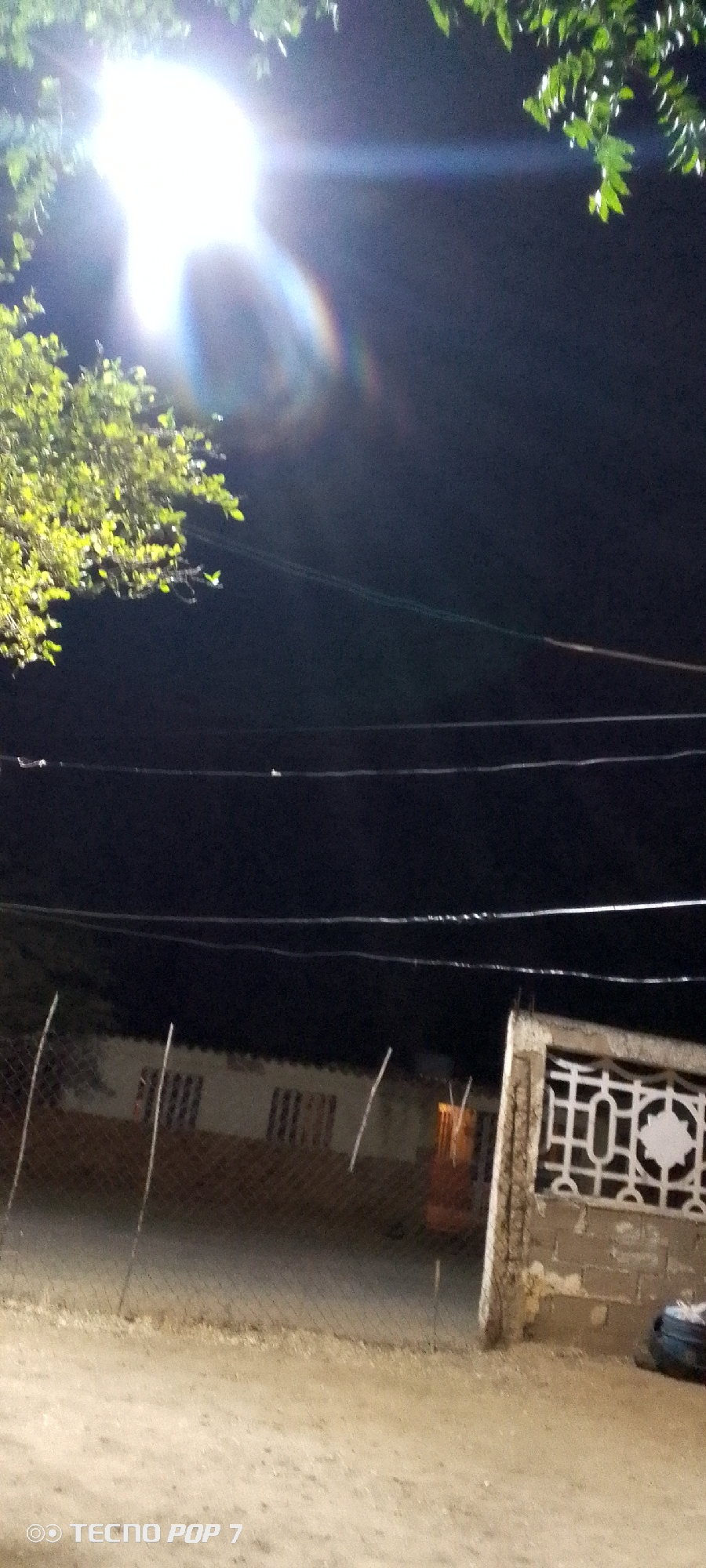 The image, captured at night, shows an outdoor scene in Altagracia, Venezuela. Bright illumination, likely from a streetlight, dominates the upper left, causing lens flare. A tree with green leaves is partially visible near the light. Power lines stretch across the upper portion of the frame. In the background, a building with visible windows is partially obscured by a chain-link fence. A section of decorative concrete wall is present on the right side of the frame. The foreground consists of a dirt ground. "TECNO POP 7" is visible in the bottom left corner.