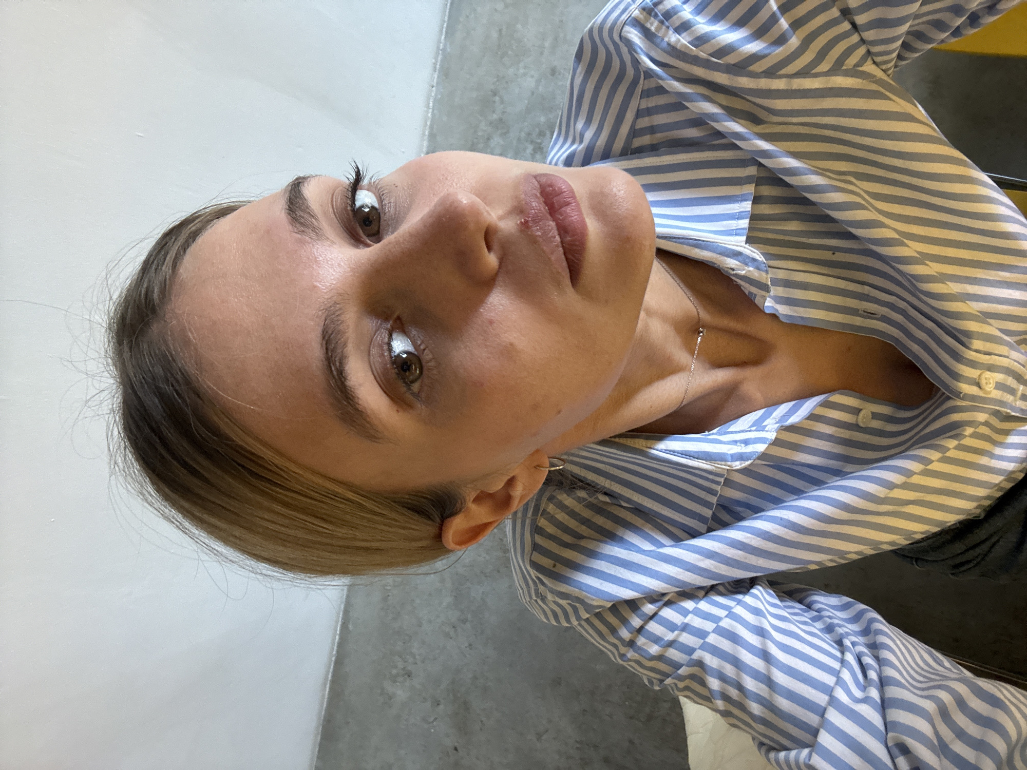 A close-up, rotated portrait shot features a person, likely female, gazing slightly upwards and to the right. The individual has light brown or blonde hair, styled away from the face, and prominent dark eyebrows framing brown eyes with long, defined lashes. The lips appear full, with a visible blemish or lesion on the upper lip. The skin appears clear, with some natural shine on the forehead and nose.
The person is wearing a blue and white striped long-sleeve button-down shirt, which covers the upper body. A thin silver chain necklace with a small, shiny pendant is visible around the neck. A small, delicate hoop earring is present in the left ear.
The background consists of a plain white wall on the left side and a grey, concrete-like floor or surface on the right and bottom. The lighting appears artificial, suggesting an indoor setting. There are no visible activities other than posing for the photograph. The expression on the person's face is neutral. No text is visible in the image.
