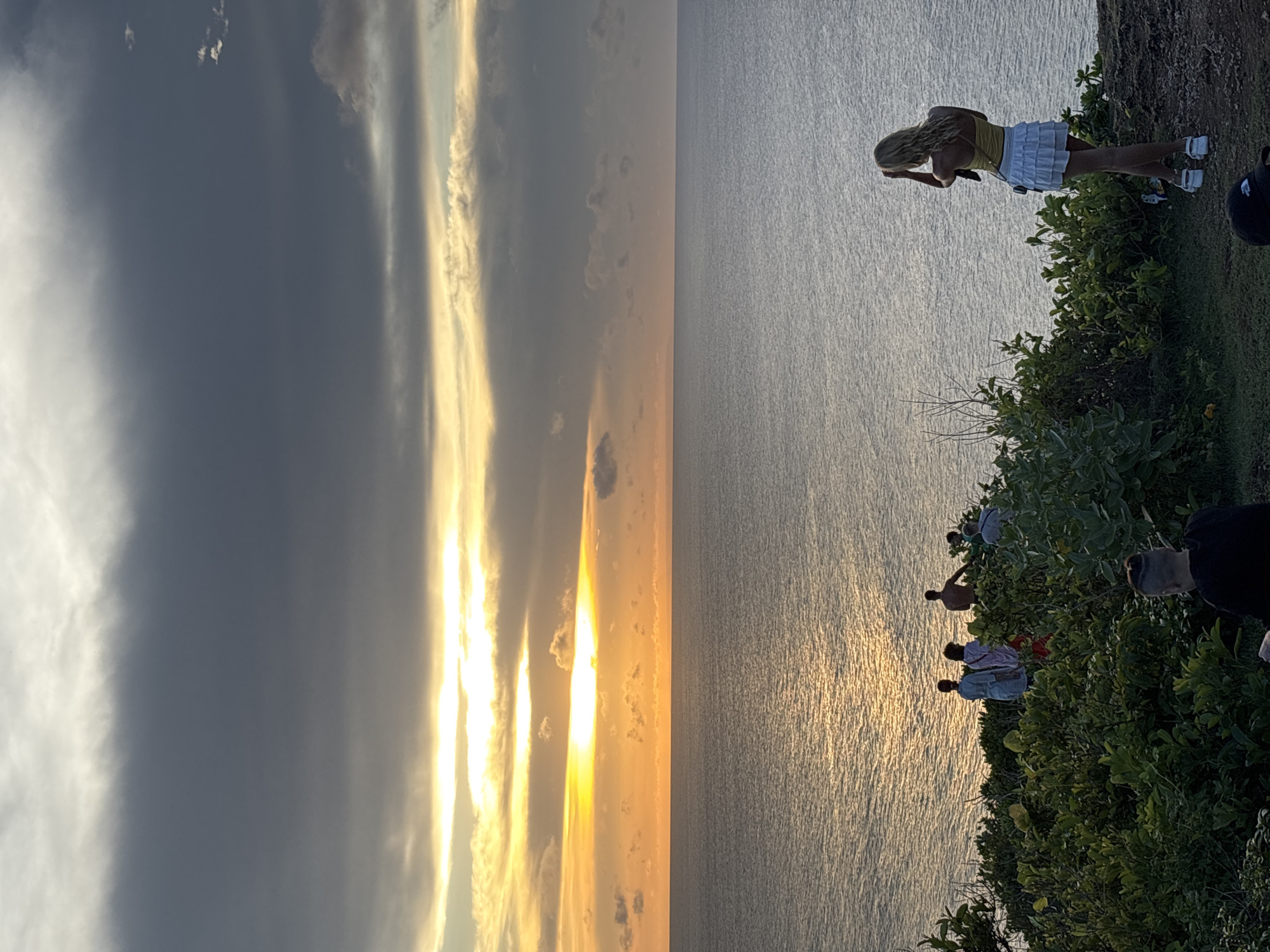 The image shows a cliffside view at sunset in Uluwatu, Indonesia. The left side of the image shows a sky with dark and light clouds illuminated by a vibrant orange and yellow sunset. To the right of the sky is the ocean reflecting the sunlight. On the right side of the image, people are standing on the edge of a cliff covered with vegetation. A blonde woman in a yellow tank top and white skirt is leaning over the edge of the cliff. Other people are standing nearby, some wearing light-colored clothing.