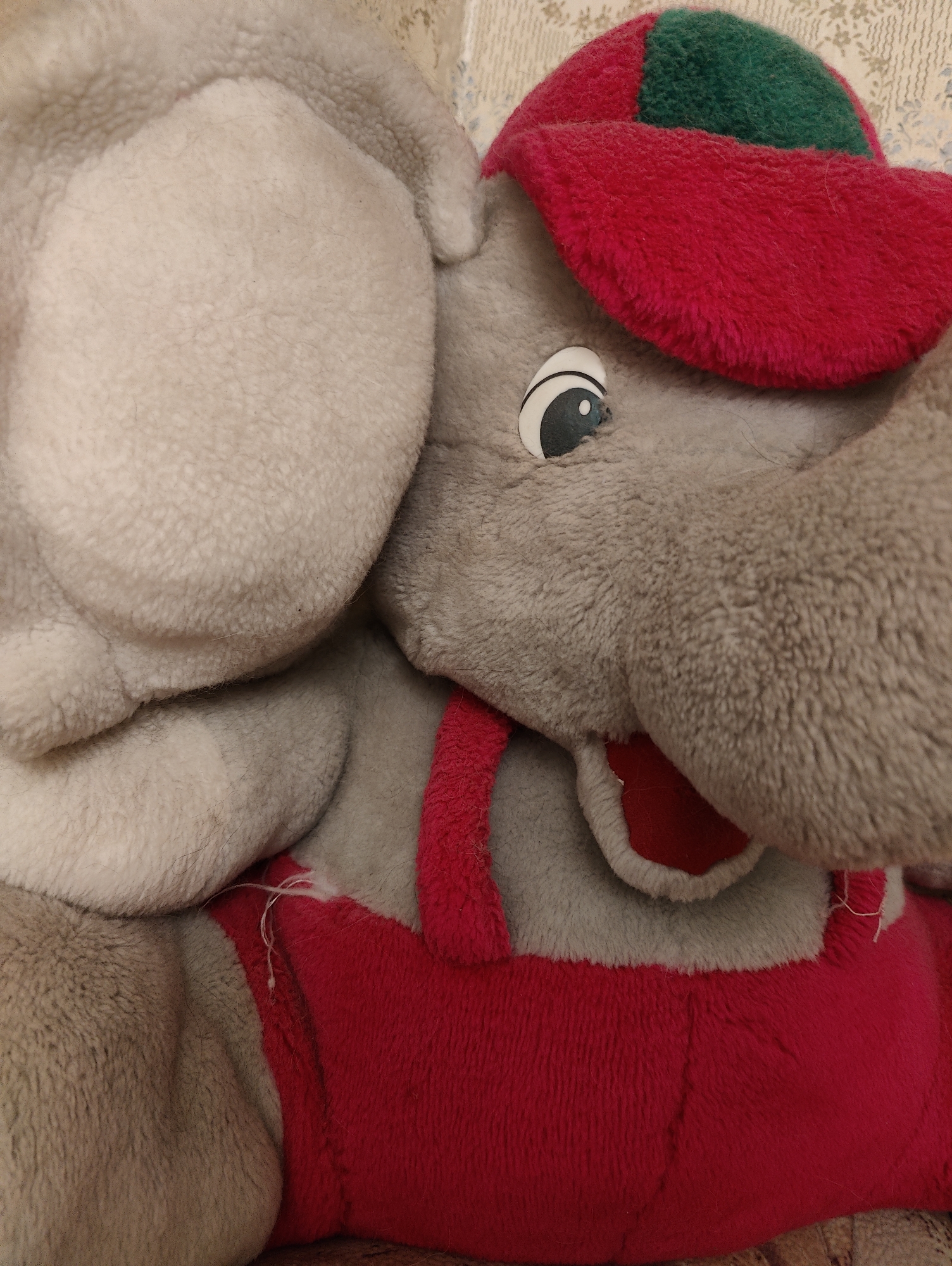 A close-up, eye-level shot features a plush toy elephant, primarily gray, wearing a red hat with a green patch and red overalls. The elephant's large head and upper torso dominate the frame, with its left ear prominently displayed on the left side. The plush material of the elephant's body and ears appears soft and textured, varying from a lighter, almost white-gray on the ears to a darker, more typical gray on the face and trunk.
The elephant has a large, cartoonish eye visible on its right side, consisting of a white base with a black pupil and a small white reflective crescent. A red hat covers the top of its head, with a distinct green section on the crown. Below its head, a portion of its red overalls with two thin straps is visible. The plush fabric of the red clothing also has a soft, slightly matted appearance, and a few loose white threads are noticeable near one of the straps, suggesting wear. A hint of the elephant's open mouth, showing red fabric inside, can be seen beneath its trunk.
The background is softly blurred but appears to be a light-colored, patterned fabric or wallpaper, possibly beige or cream with a subtle floral or decorative design. There is no discernible activity taking place, and no text is visible in the image. The lighting is even, suggesting an indoor setting.