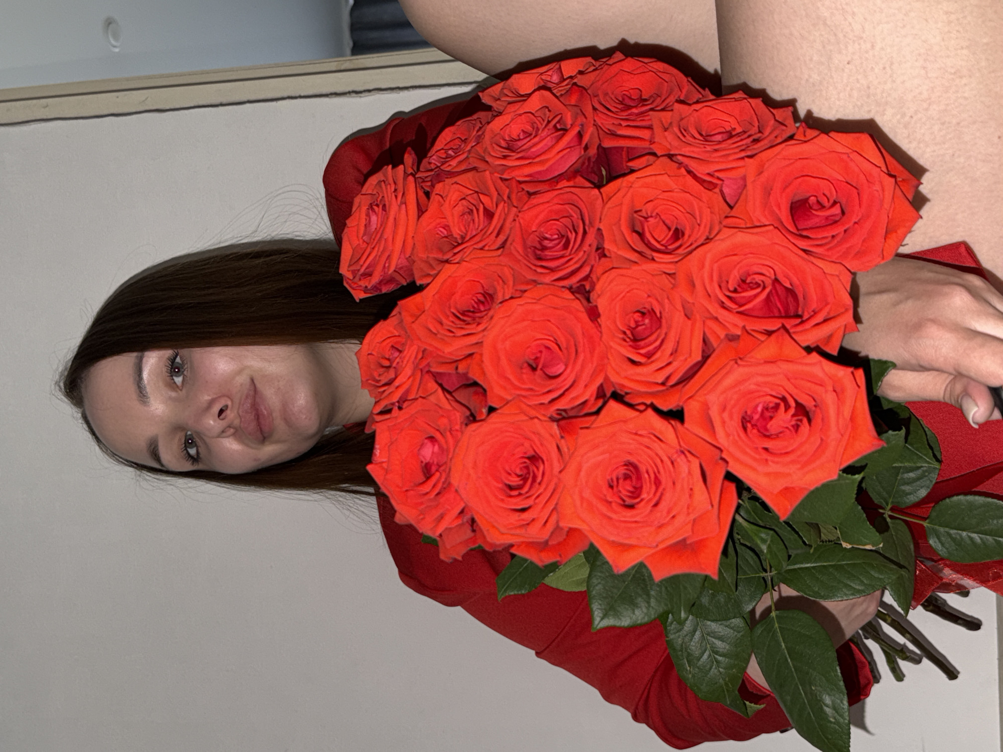 A young woman with long brown hair holds a large bouquet of red roses. She wears a red garment and is positioned in front of a light-colored wall. The location is inferred to be Chaikovsky, Russia.