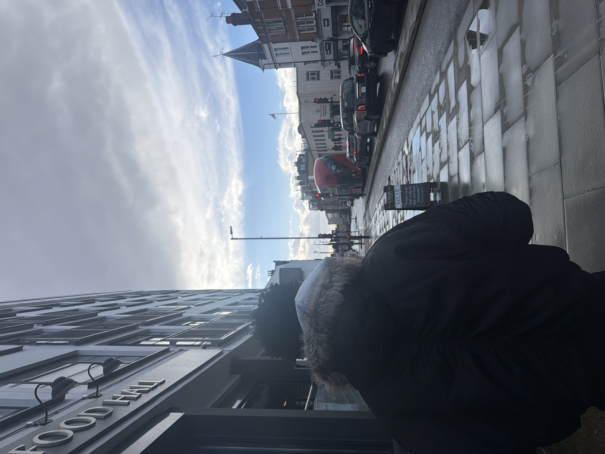 The image depicts a street scene in London, United Kingdom, captured from behind an individual on a wet sidewalk. The foreground features a person wearing a dark, potentially black, ruched-back jacket with a grey fur-lined hood, seen from the rear. The sidewalk is paved with large, reflective grey slabs, indicating wet conditions. To the left of the person, a partially obscured A-frame sign displays text including "COOK - RELAX" and "Frozen meals that really do look and taste homemade."
To the immediate left, a multi-story building with numerous windows features a sign partially reading "FOOD HALL" in large, light-colored letters. The asphalt road, also wet, shows traffic moving away from the viewer. Vehicles include a red double-decker bus in the left lane, followed by a black London taxi and several other cars. Traffic lights further down the street are illuminated red.
Across the street, a line of buildings is visible. A prominent white-faced building features a dark conical spire on its roof, above which a Union Jack flag flies from a flagpole. Other commercial signs, including "Dearden" and a partial "CO-OP", are visible on adjacent structures. The sky is partially cloudy with patches of blue visible, suggesting dynamic weather conditions following rain.