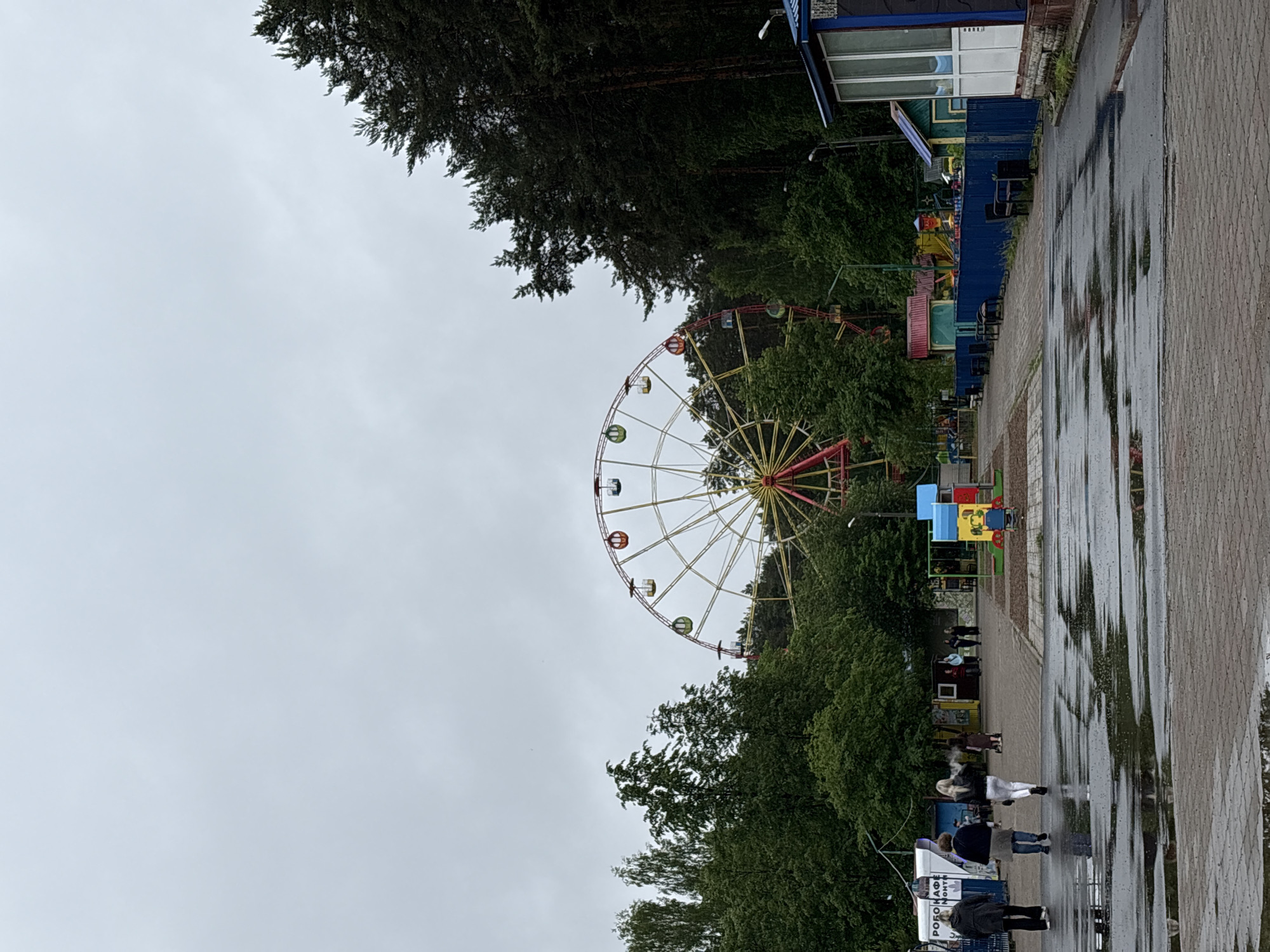 This image depicts a scene from an outdoor amusement park, likely Gorky Park in Perm, Russia, captured on an overcast day. The foreground shows a wide, paved pathway, possibly constructed from interlocking pavers, heavily marked with numerous puddles, indicative of recent rainfall.
On the left side of the pathway, several individuals are walking away from the camera. Prominently, three women are visible closer to the viewer: one in a dark jacket and dark trousers, another in a dark jacket and blue jeans, and a third with blond hair wearing a white jacket and white trousers, from whom a visible plume of white vapor emanates. Further along the path, additional pedestrians are present. To the right, a low, blue metal fence runs parallel to the path, behind which black benches and small, modest buildings with varied roof colors are situated. A more substantial building with a blue roof and a white and glass facade is partially visible on the far right edge of the frame.
The midground is characterized by a dense expanse of green trees, predominantly deciduous, though some darker coniferous trees are visible on the right. Towering above the tree line in the center-right is a large Ferris wheel, featuring a distinct yellow and red structural framework and multicolored passenger cabins in shades of red, yellow, green, blue, and orange. To the left of the path, a white kiosk or building with "РОБОКАФЕ" (Robocafe) and "Монт" written in Cyrillic is visible. A colorful playground area, featuring blue, yellow, and red equipment, is visible on the right side of the pathway, beyond the blue fence, with a child in blue clothing near the equipment.
The background consists of a continuation of dense tree cover, with the upper portion of the Ferris wheel prominent against the uniformly grey, cloudy sky.