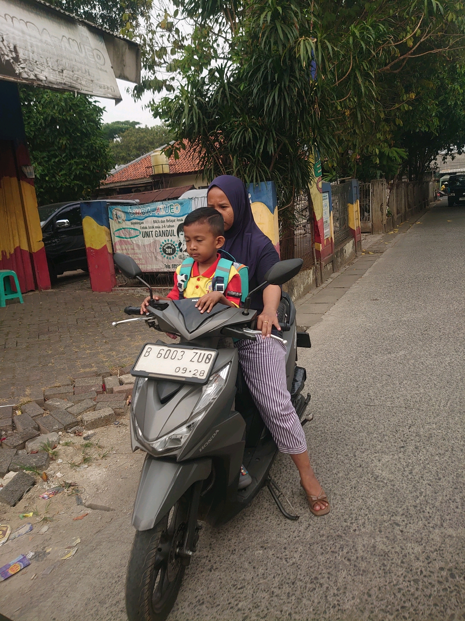 A medium shot captures an Indonesian woman wearing a purple hijab and striped pants, seated on a gray scooter. She is holding the handlebars and has her right hand on them. A young boy, likely her son, is sitting in front of her, also on the scooter. He is wearing a yellow and red shirt with a blue backpack and a small backpack. The scooter's license plate reads "B 6003 ZUB 09-28". The background shows a street scene in Depok, Indonesia, with buildings, trees, and a fence. The weather appears overcast.