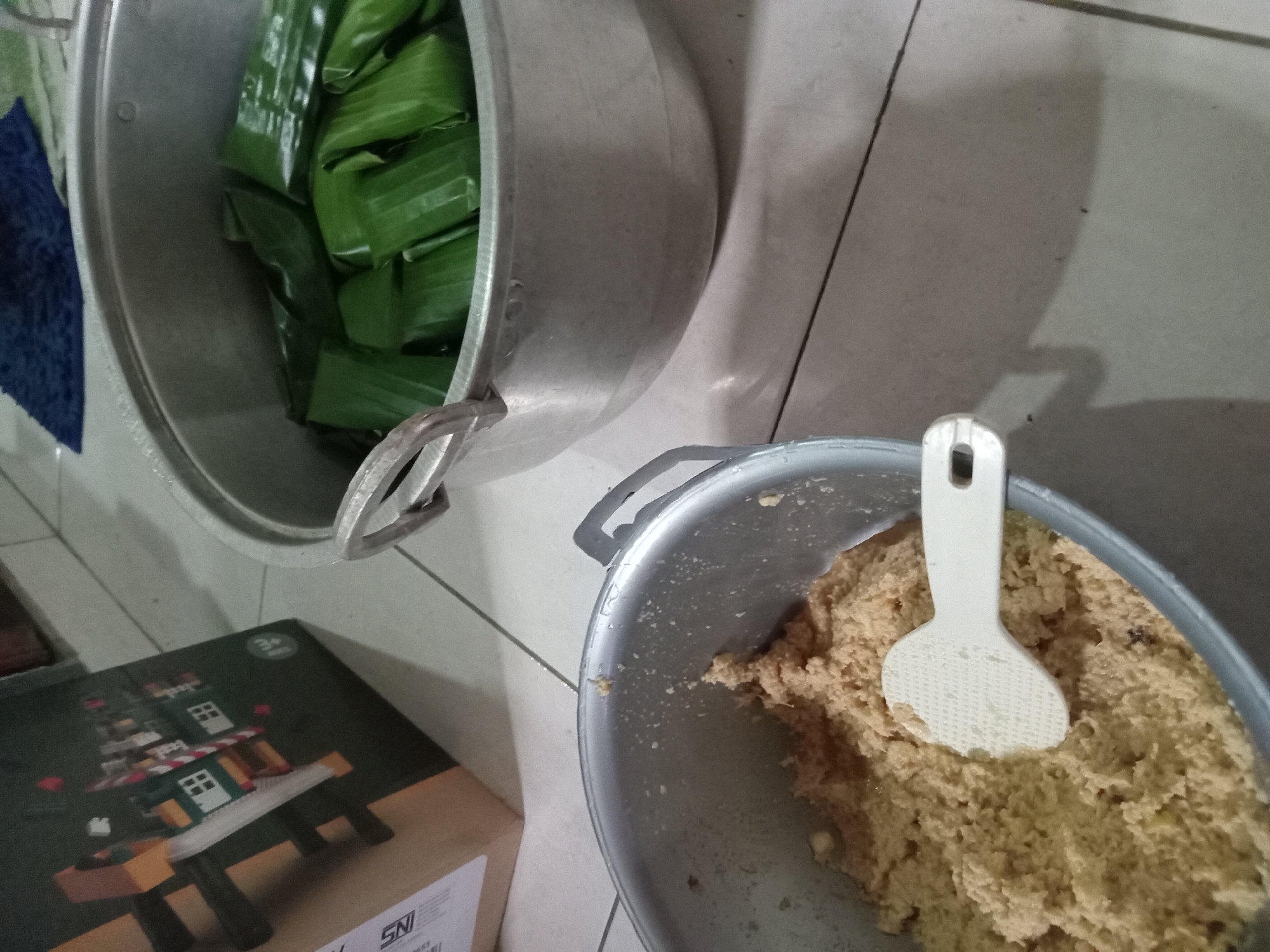 A top-down shot shows a cooking scene with two aluminum pots on a tiled floor. In the foreground, a large aluminum pot is filled with a coarse, light brown mixture, possibly rice or a dough, with a white plastic rice paddle resting inside. To the right, a larger aluminum pot contains numerous green, rectangular items wrapped in what appears to be banana leaves, suggesting food being prepared for steaming or cooking. Behind the pots, a cardboard box with graphics of a building structure is partially visible, and a dark blue fabric is visible in the background. The overall scene suggests food preparation in a domestic setting in Claket, Indonesia.