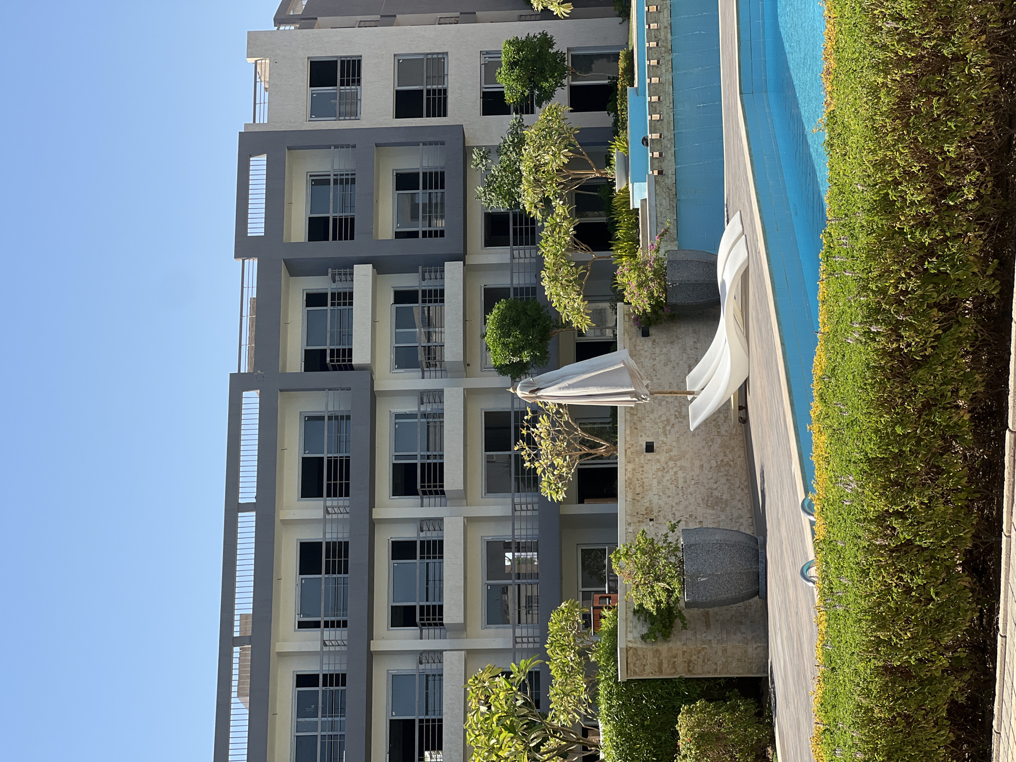 The image shows a modern apartment building with multiple floors and balconies, set against a clear blue sky. In the foreground, there is a swimming pool with blue water and a stone-tiled edge. Two white lounge chairs are placed on a wooden deck beside the pool, with a folded umbrella leaning against one of them. Green bushes and small trees are planted around the pool area, and a large grey planter is visible on the left. The building has a contemporary design with white and grey facades and numerous windows and glass doors. The overall scene suggests a resort or residential complex in a warm climate. The location is identified as Unknown, Egypt.