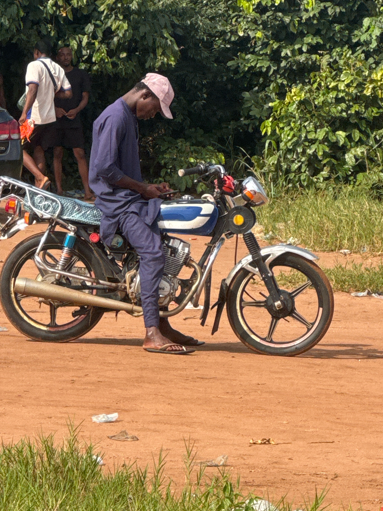 A man wearing a dark blue traditional outfit and a pink cap is seated on a blue and white motorcycle. He is looking down at his mobile phone. The motorcycle has a silver exhaust pipe and a multi-spoke wheel. In the background, there are several people standing near a car. The scene is outdoors, with lush green foliage and trees behind the motorcycle. The ground is covered in reddish-brown dirt. There are also some dry leaves and white debris scattered on the ground. The image appears to be taken in Ihiagwa, Nigeria, given the context of the request.