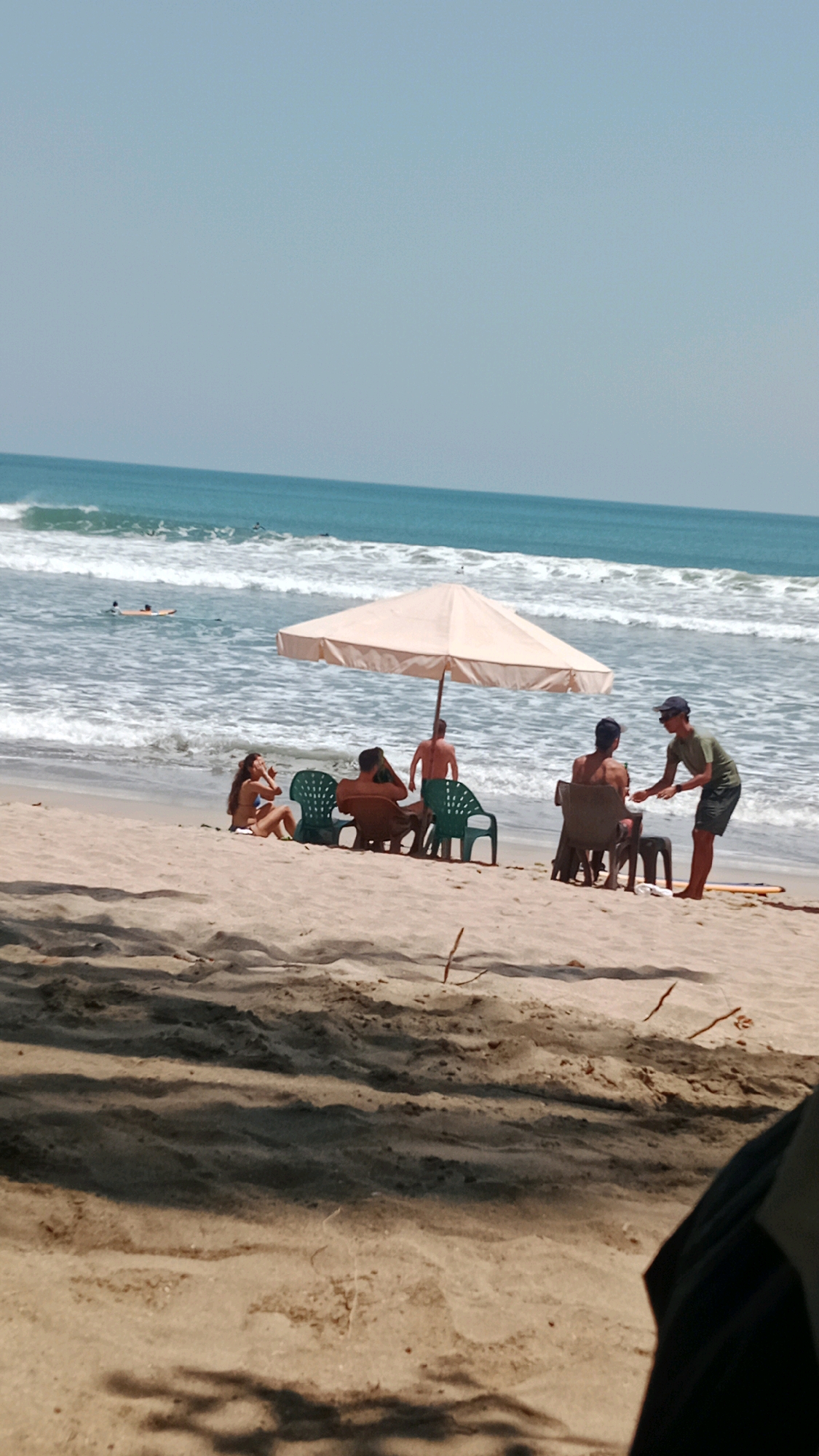 In Kuta, Indonesia, a group of people are seen enjoying a day at the beach. The scene captures a relaxed atmosphere with individuals seated on beach chairs under a large, light-colored umbrella. Two people are facing each other, engaged in conversation, while another person stands nearby, seemingly interacting with them. In the background, the ocean stretches out with gentle waves, and several surfers are visible in the water, enjoying the ocean's offerings. The sandy beach, characteristic of Kuta, provides a warm and inviting setting for this beach activity. The overall scene portrays a typical leisure day at one of Bali's most famous beaches.