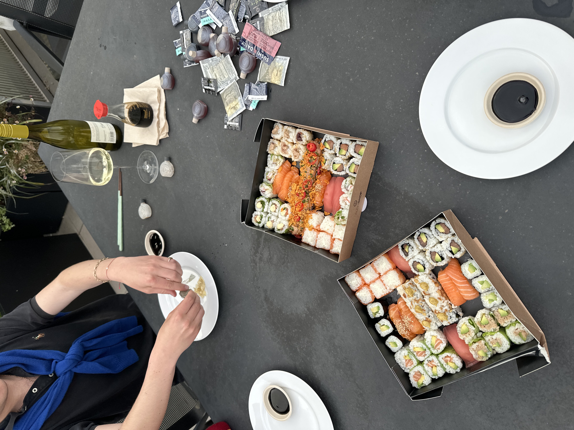 The image presents an overhead view of an outdoor sushi meal enjoyed on a dark grey or black table, likely a patio or balcony. One person is partially visible in the bottom left, wearing a black shirt with a blue sweater tied around their shoulders. Their hands are actively squeezing a small packet onto a white plate, which already contains ginger and what appears to be wasabi.
Two large cardboard boxes are filled with an assortment of sushi, including various maki rolls, nigiri (salmon, tuna), and sashimi, featuring ingredients like salmon, tuna, avocado, and diverse toppings. Both boxes display the "SUSHISHOP" logo.
Scattered across the table are numerous small packets of soy sauce, some labeled "SUSHI WASA" and "SOY SAUCE DAILY," alongside several small, single-serving plastic bottles of soy sauce. A green bottle of white wine, possibly branded "La Vendange," stands beside a wine glass containing wine, with another empty wine glass nearby. A pair of green chopsticks rests on the table. Three white plates are visible; two hold small circular containers filled with dark soy sauce, while the third is being prepared by the person.
The scene suggests a casual daytime meal, possibly a takeout or delivery experience, set in a relaxed outdoor environment in London, United Kingdom.