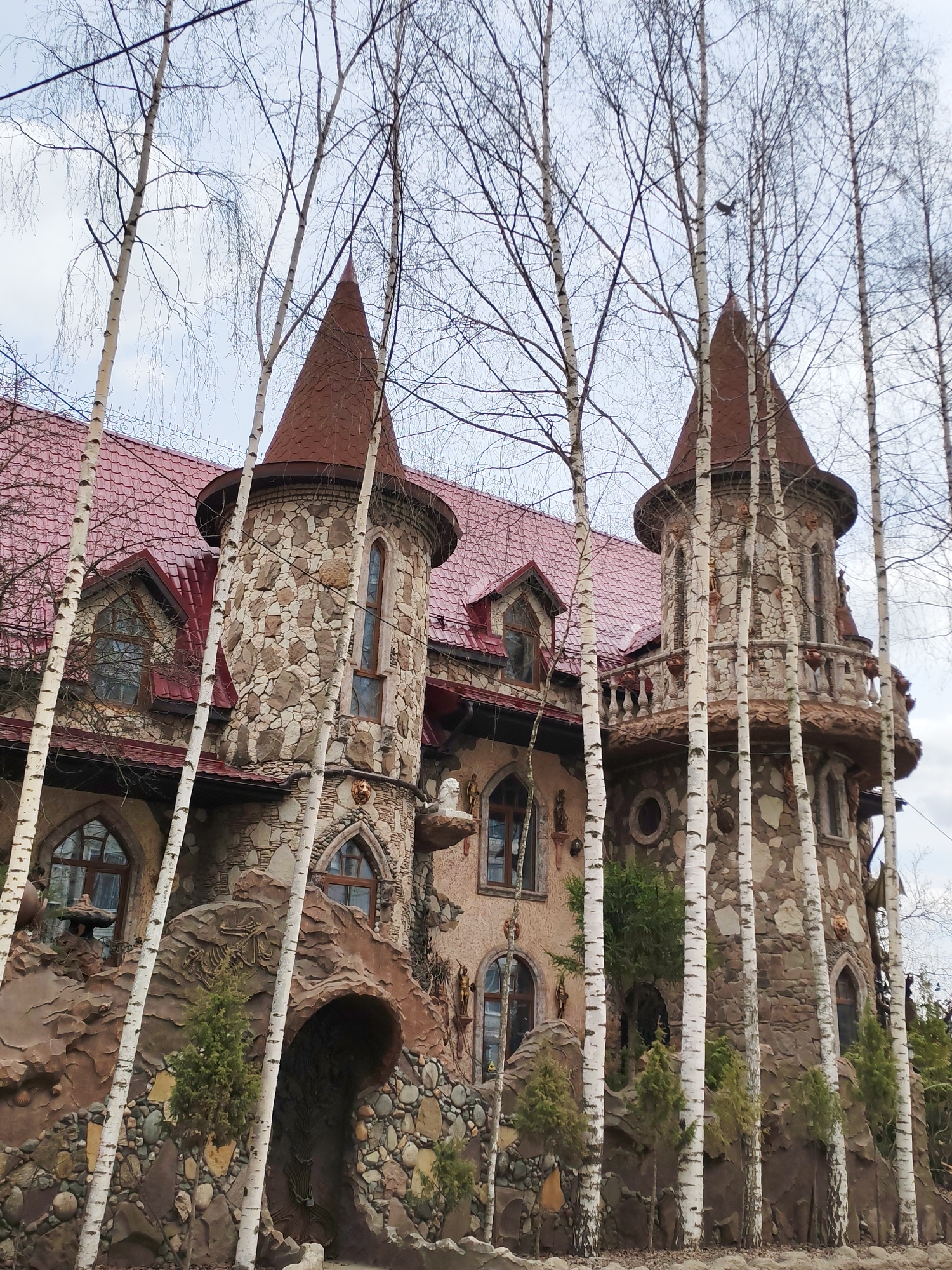 The image shows an elaborate, fairytale-like castle with two prominent towers, located in Izhevsk, Russia. The building is constructed primarily of stone, with a red-tiled roof. The towers feature conical roofs, windows, and decorative elements. The exterior is adorned with sculptures and other embellishments. In the foreground, several birch trees with bare branches partially obscure the view of the castle. A stone archway leads into what appears to be an entrance. Small evergreen shrubs are planted near the base of the building. The sky is overcast.