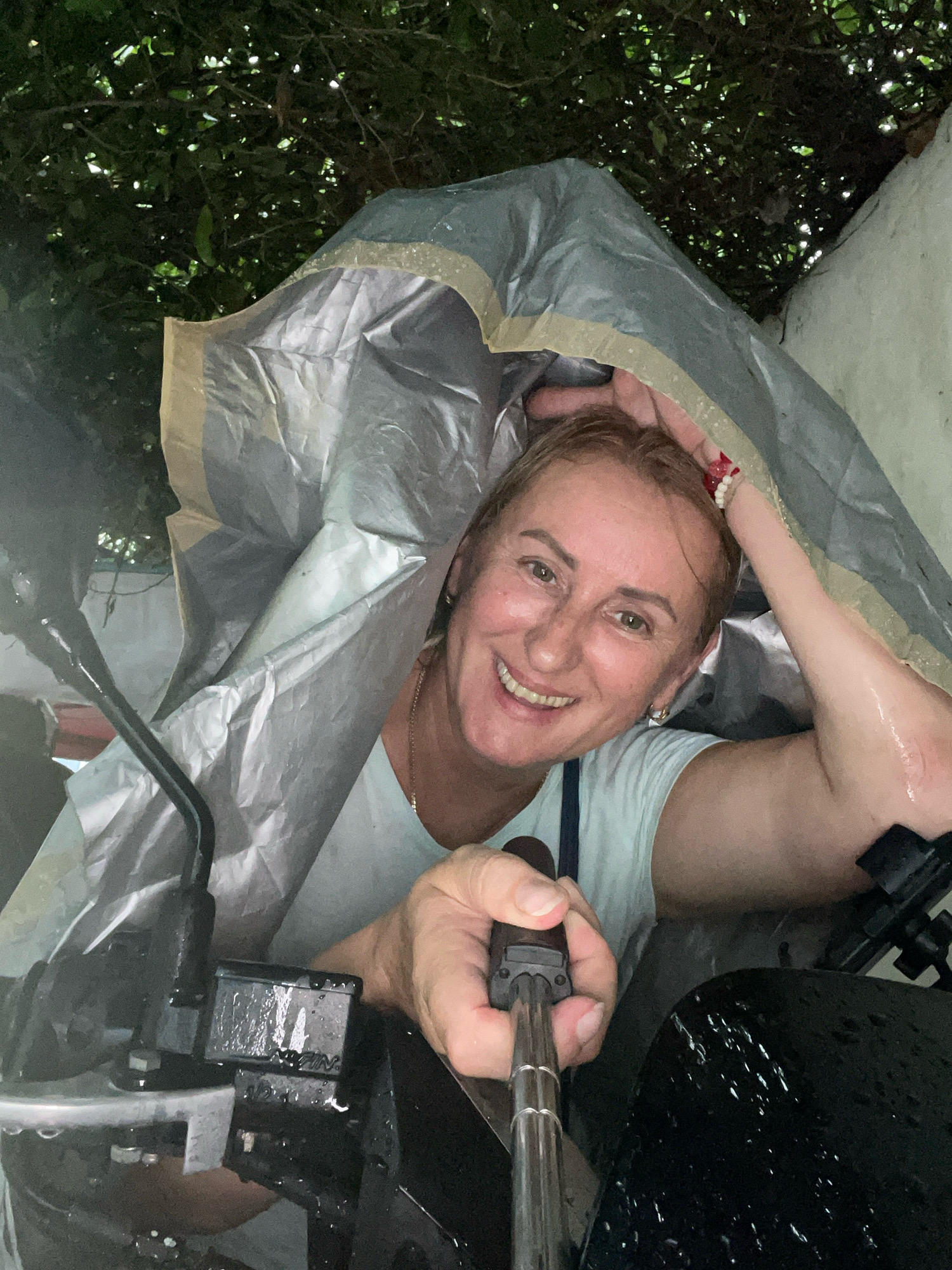 In Pattaya City, Thailand, a woman is taking a selfie with a motorcycle under a tarp during a rainstorm. The woman, wearing a light blue t-shirt, is smiling at the camera, holding a selfie stick. She is partially covered by a silver and tan tarp, with parts of the motorcycle, including the handlebars and a control panel, visible and wet. The environment appears to be outdoors, with lush green foliage visible in the background and a light-colored, possibly stucco, wall on the right. Droplets of water are visible on the tarp, the motorcycle, and the woman's arm.