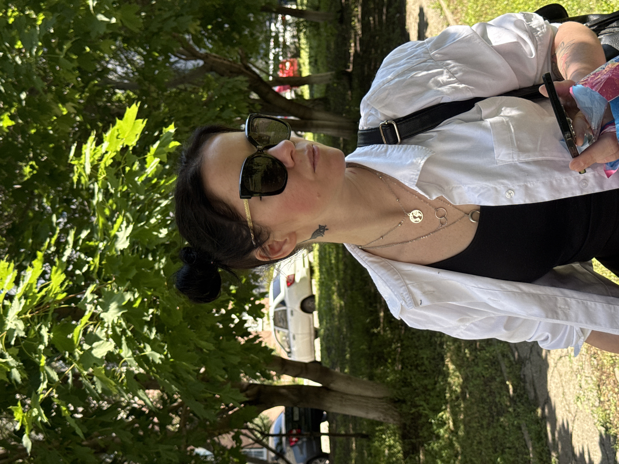 In the city of Unknown, Unknown, a woman stands in front of a backdrop of trees and parked cars. She is facing the right of the frame, wearing a white button-up shirt over a black tank top. Her hair is pulled up in a bun, and she has on large, dark sunglasses. A tattoo is visible on her neck. She is adorned with multiple necklaces, and a black backpack is slung over her shoulder. In her right hand, she holds a smartphone and a small, colorful bag. The ground is paved with concrete, with grass and trees surrounding the area. The scene appears to be set during the daytime, with sunlight filtering through the leaves.