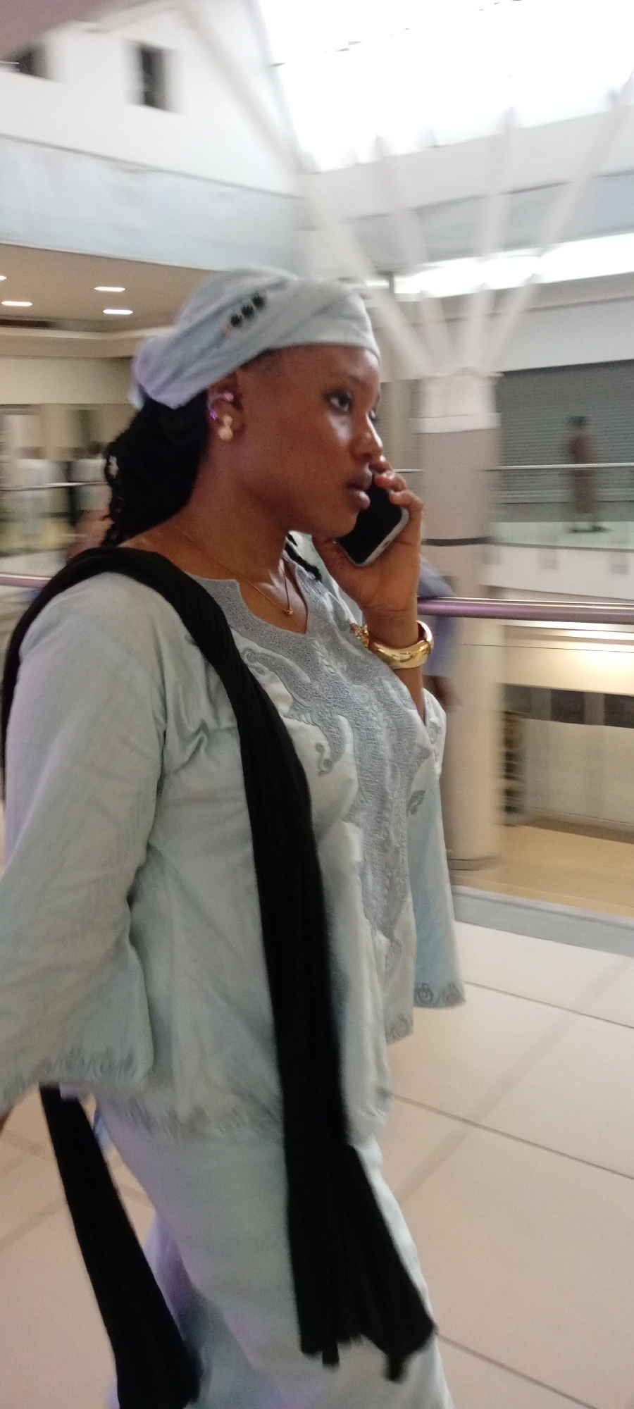 In Gandu Albasa, Nigeria, a woman in a light blue headscarf and a matching, intricately embroidered tunic walks while talking on a mobile phone. She wears large, pearl-like earrings and a gold bracelet. A black scarf is draped over her shoulder and chest. The background suggests an indoor public space, possibly a mall or airport, with blurred architectural features and what appear to be shops or kiosks.