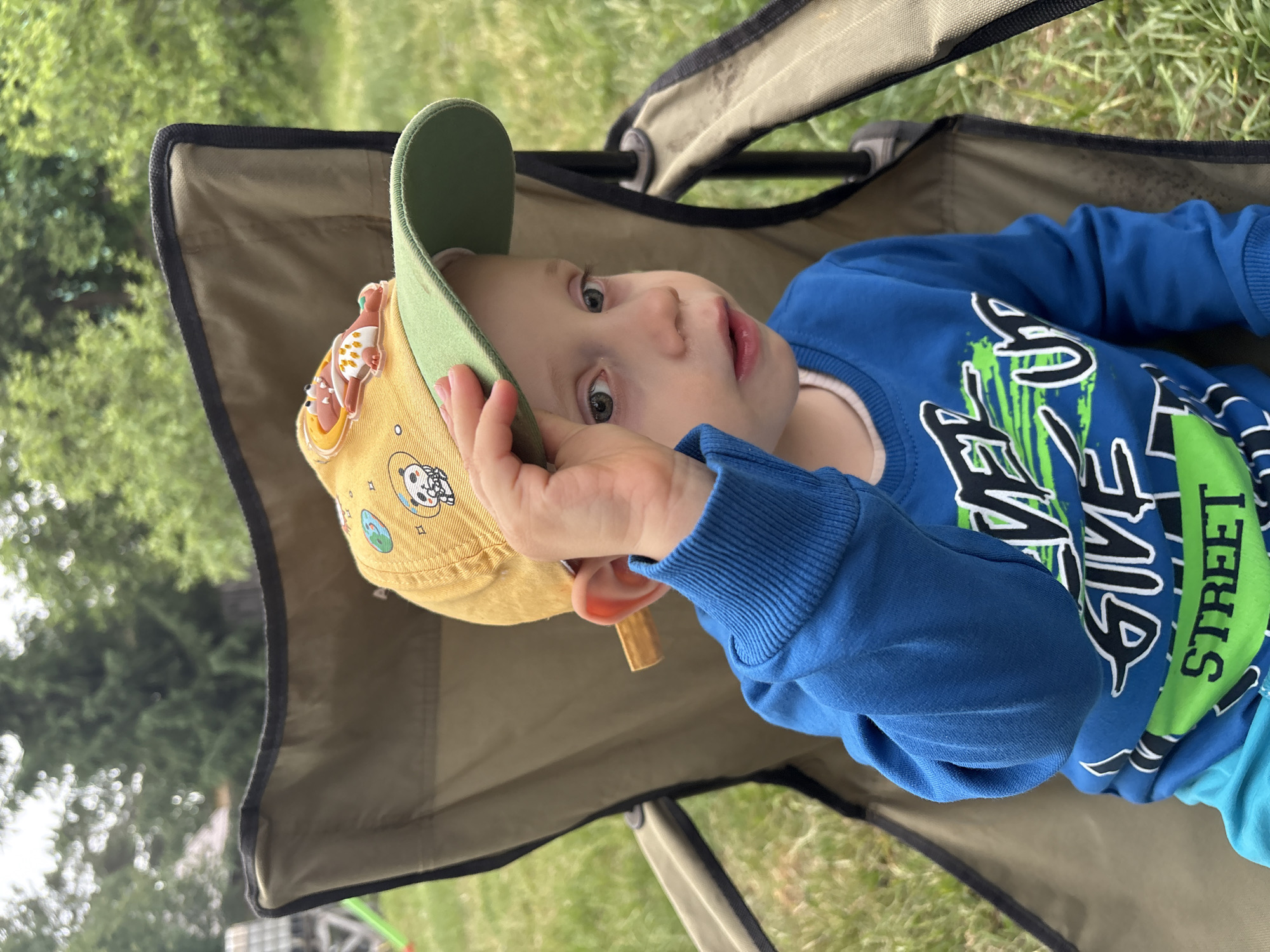 A child wearing a yellow and green hat with cartoon characters is reclining in a khaki and black folding camping chair outdoors. The child is wearing a blue long-sleeved shirt with black and green graphics. The background shows green foliage. The location is unknown, and there is no verifiable indication that the photo was taken in Chaikovsky, Russia.