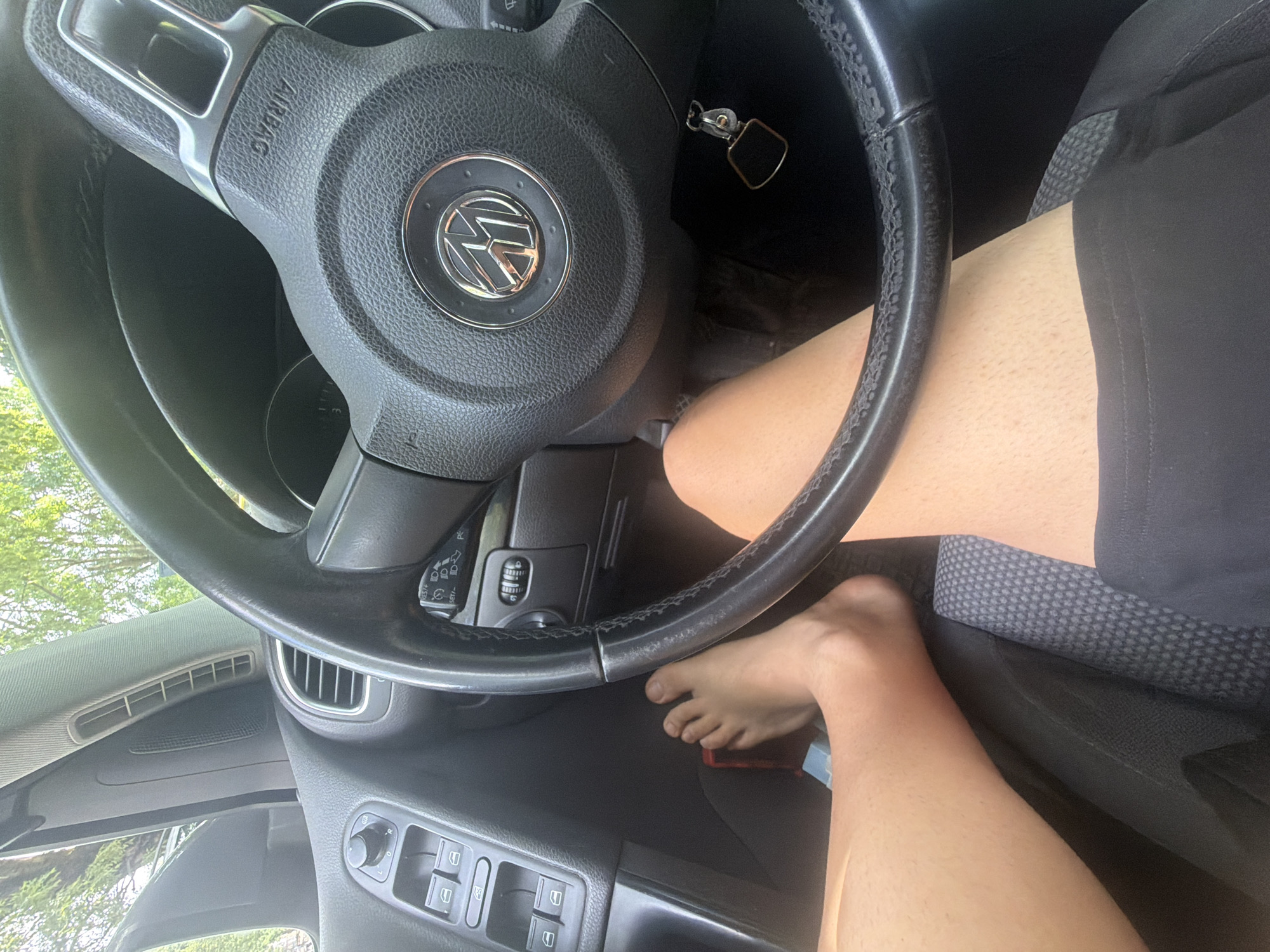 The image displays the interior of a car, focusing on the driver's perspective, likely in Iskar, Bulgaria. The central subject is the steering wheel, bearing a prominent Volkswagen logo and the word "AIRBAG." A person's bare legs and feet are visible; one foot rests on the car floor, while the other leg is bent, with the foot positioned near the steering column. The person is wearing black shorts.
The car's ignition key is visible, hanging from the steering column. Through the car's window, bright natural light illuminates lush green foliage, indicating an outdoor setting during daytime with clear weather.
Other visible car components include air vents on the dashboard, a door panel with window controls, and a portion of the driver's seat upholstery. The left stalk behind the steering wheel shows icons for lights and wipers, along with "SET/-" and "RES/+" text, likely for cruise control functions. Faint numbers "1" and "3" are discernible on a control unit located below the left air vent. A small, rectangular object, possibly a phone, is partially visible near the driver's feet. The steering wheel's leather trim shows signs of wear. The scene suggests the car is stationary or parked.