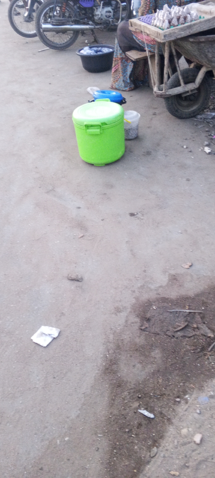 The image shows a street scene in Zango-ndaji, Nigeria. A green cooler and a white bucket are on the left side of the frame. Next to them is a blue lid. Further away are 2 motorcycles. Further back is a wheel barrow with various containers, and next to that is a person with blue and red clothing. The ground is mostly concrete or asphalt, and there is trash on the ground. There is a small white piece of litter in the lower left of the image.