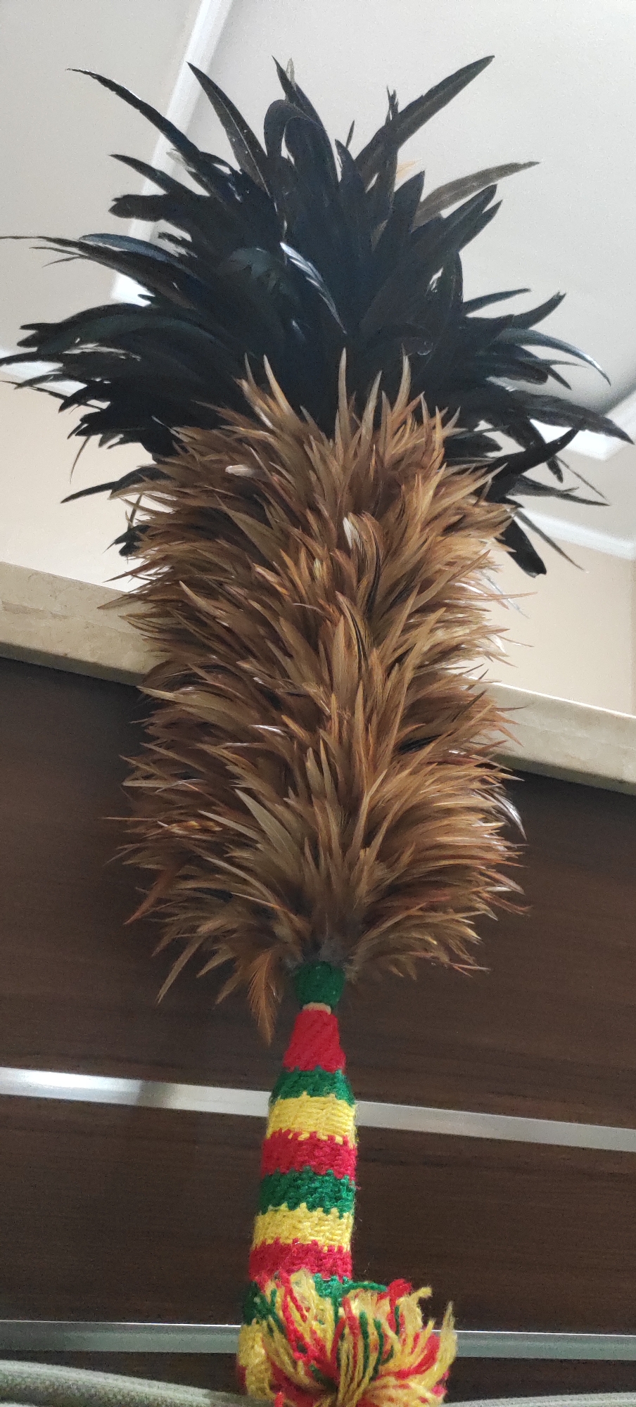 A feather duster with black and brown feathers is hanging from a shelf. The handle of the duster is decorated with colorful knitted yarn in red, yellow, and green stripes, and a tassel of red, yellow, and green yarn hangs from the bottom. The background is a plain wall. The location is Manyar, Indonesia.