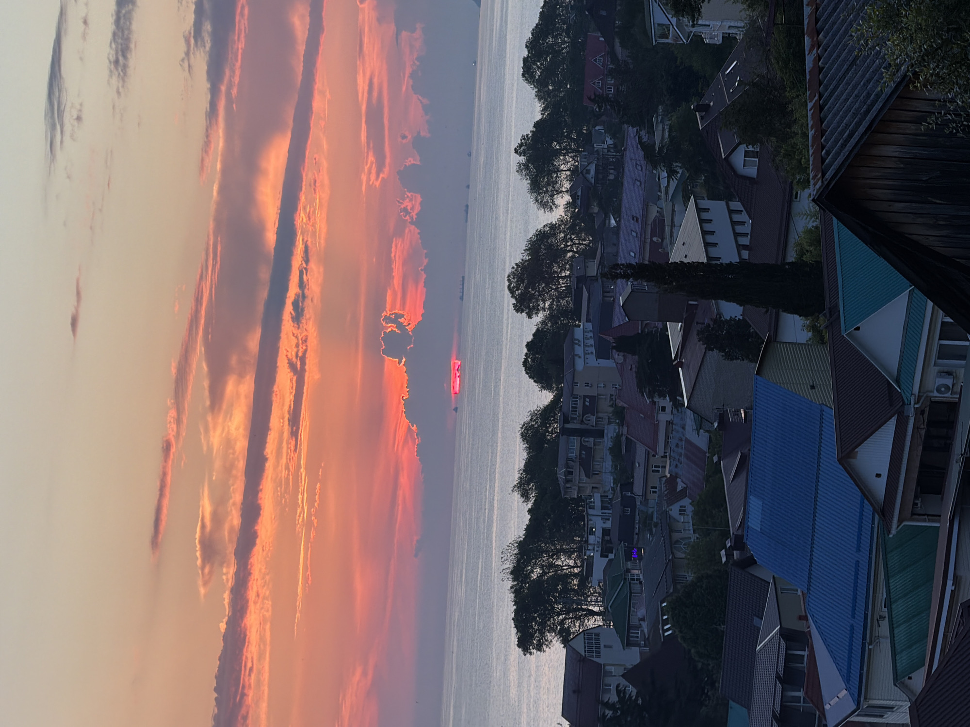 An aerial, eye-level, wide shot captures a sunset over a body of water. The sun, a bright red-pink hue, is partially obscured by clouds, creating a radiant glow. The sky is painted with warm tones of orange, pink, and yellow, with scattered clouds adding texture. The water, a calm expanse of gray-blue, stretches towards the horizon where several ships are visible as dark silhouettes. Below the water lies a densely packed residential area. Houses with a variety of roof colors, including blue, green, and brown, are clustered together, interspersed with trees. The architecture suggests a Mediterranean or similar coastal style. The overall scene is one of a serene evening, with the natural beauty of the sunset complemented by the human-made landscape.