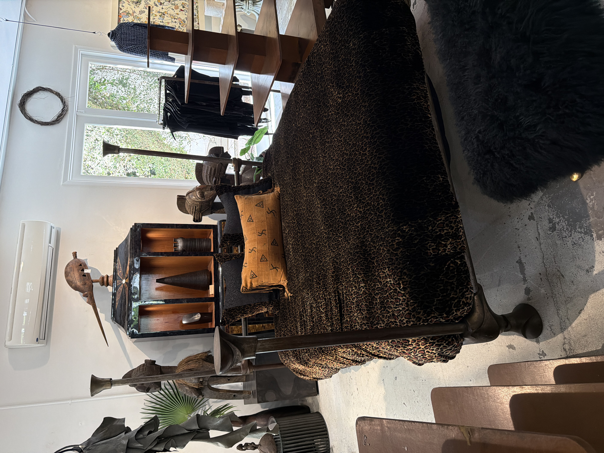 The image captures an interior scene in what appears to be a store or showroom, possibly located in Los Angeles, United States of America. The interior is characterized by an eclectic mix of furniture and decorative objects. A bed with a leopard-print cover and decorative pillows is prominent.
Alongside the bed, there is a black cabinet housing a variety of decorative objects, including vases and cones. In front of the cabinet, and around the bed, are various sculptures and other decorative pieces.
A window is visible in the background, offering a glimpse of greenery outside. There is a wreath hanging on the wall near the window. Additionally, clothing racks and shelves are positioned in the upper part of the frame. The scene is bathed in natural light, indicating daytime. The combination of objects suggests an artistic or design-focused space.