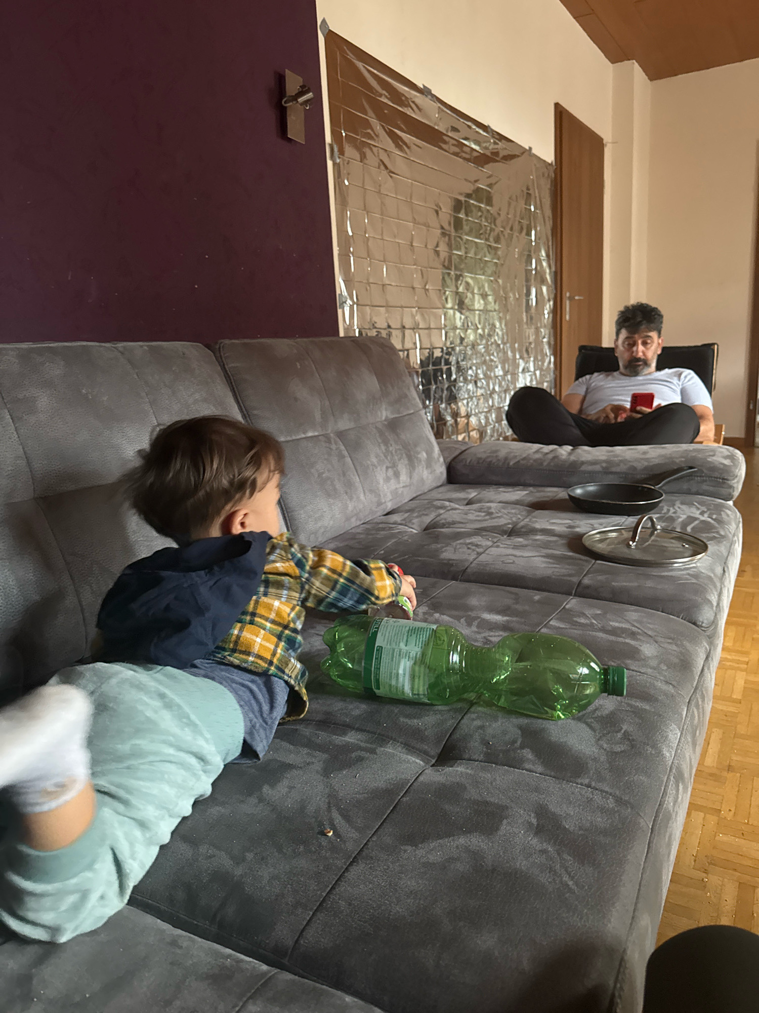 A young child in a plaid shirt and light blue pants lies on a dark gray couch, facing away from the camera and to the left. A man in a light blue t-shirt and dark pants sits in a chair in the background, looking at his red smartphone. A silver foil emergency blanket hangs on the wall behind him. On the couch in front of the man, there is a small black frying pan and a glass lid. A green plastic water bottle lies on the couch near the child. The room has hardwood floors and wooden doors. The scene appears to be indoors, likely a living room, in Überherrn, Germany.