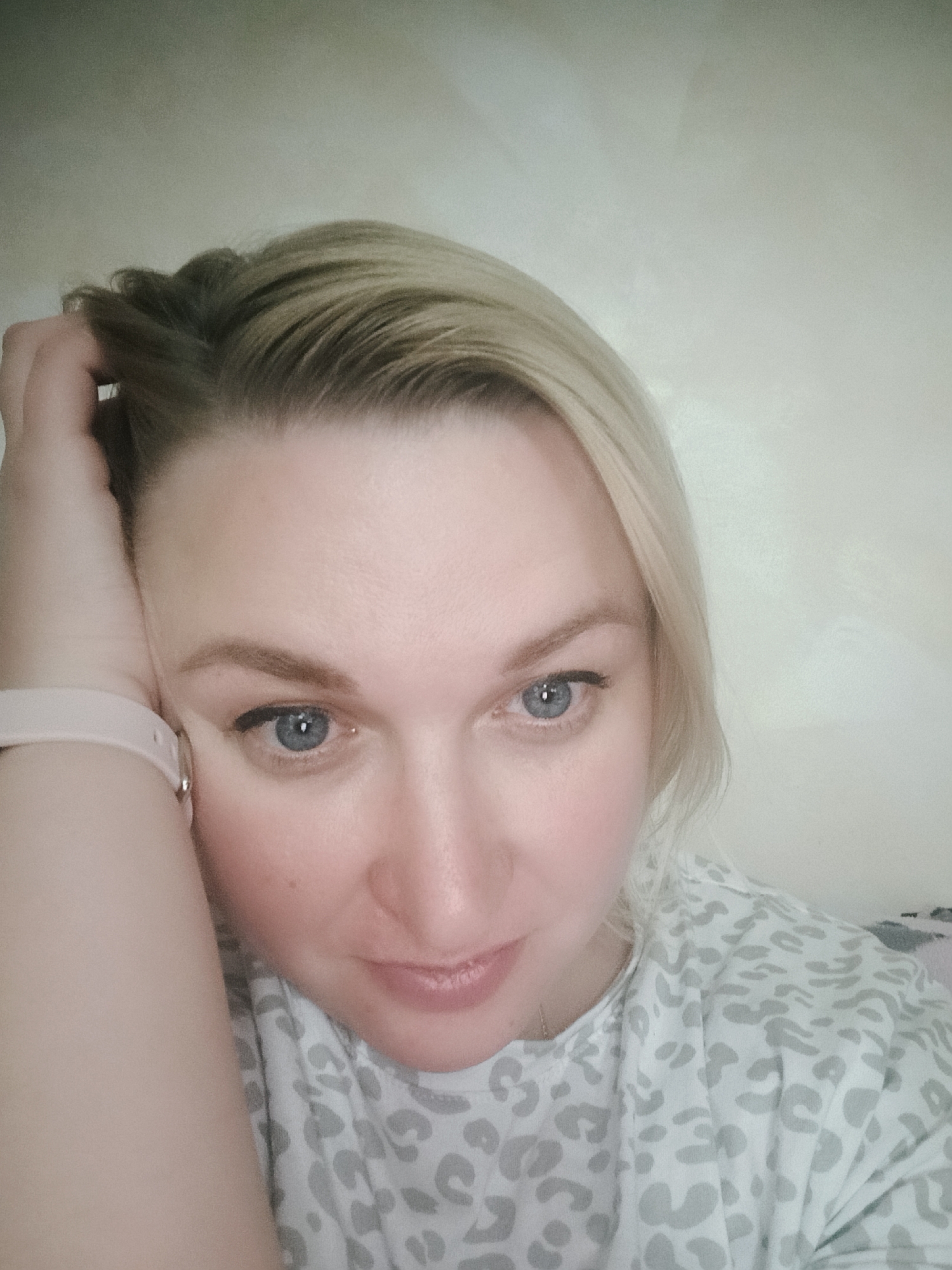 A close-up selfie of a blonde woman with light blue eyes. She has her hand on her head, and is wearing a light pink watch. She is wearing a white shirt with gray leopard print. The location is difficult to determine and cannot be determined from the image alone.