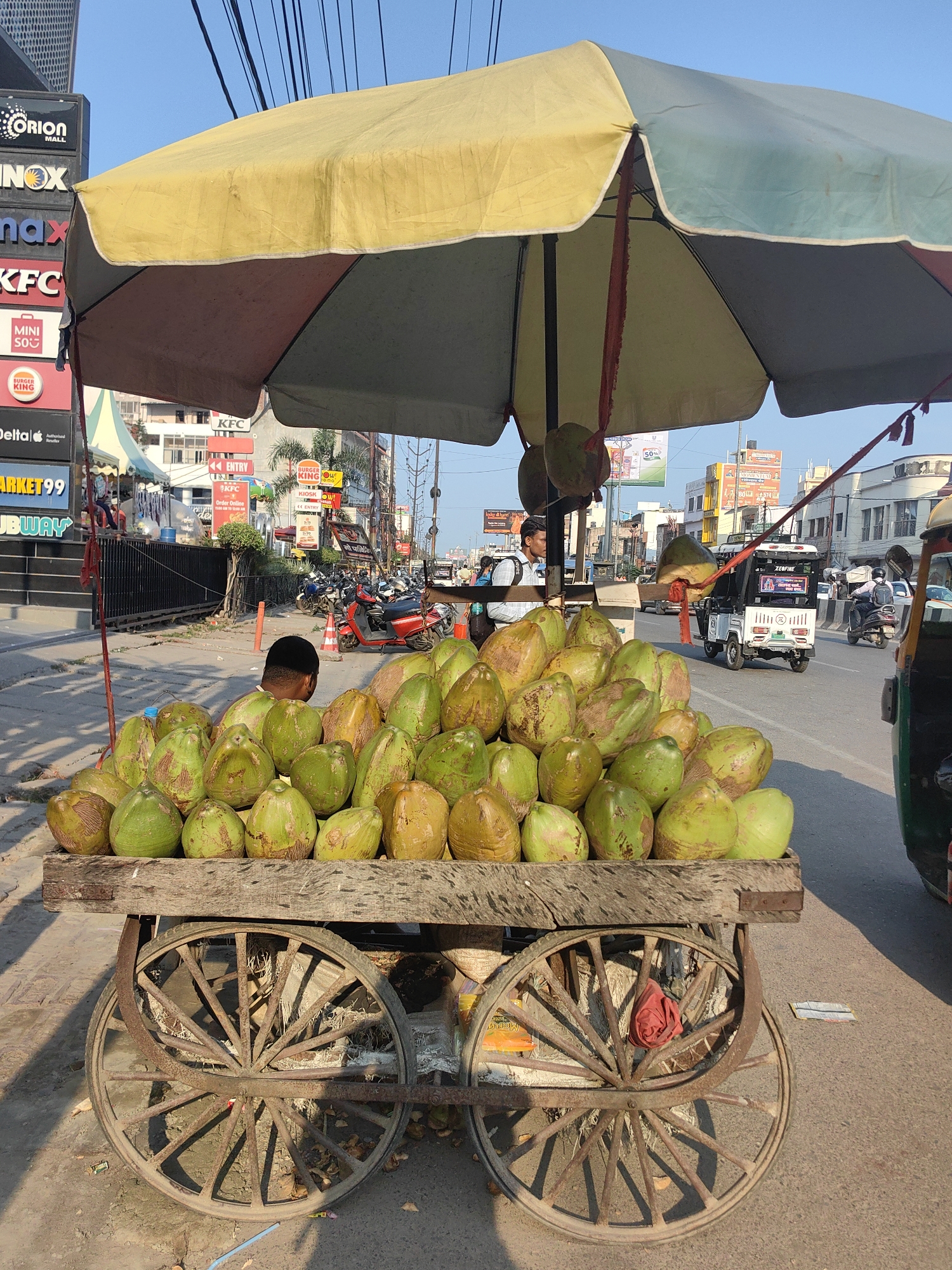 A street vendor's cart laden with green coconuts is positioned on a paved road in Bichhia, India. The cart, made of weathered wood and featuring large, spoked wooden wheels, is covered by a multi-colored umbrella, predominantly yellow and blue. A young male vendor, wearing a light-colored shirt and dark hair, is partially visible behind the coconuts. In the background, a busy street scene unfolds with traffic including motorcycles, a three-wheeled auto-rickshaw, and various buildings lining the road. Advertisements for businesses like Orion Mall, INOX, Max, KFC, and Subway are visible on the left side of the image. The sky is clear and blue, suggesting a sunny day.