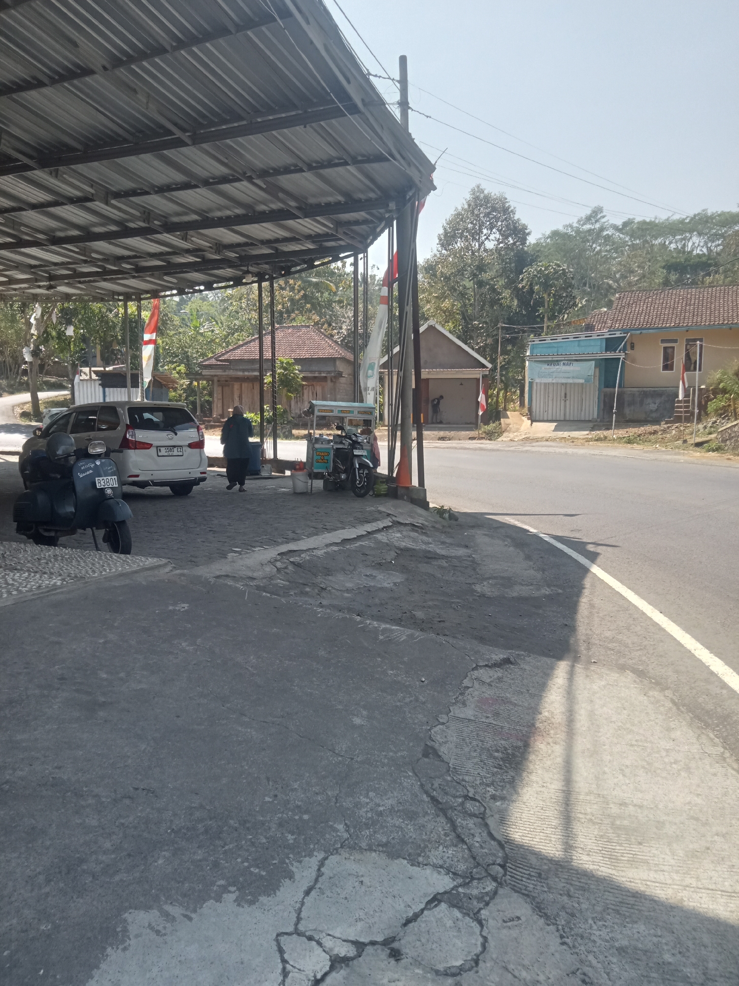 The image captures a roadside scene in Tlogosari, Indonesia. In the foreground, a cracked and worn asphalt surface transitions to a lighter concrete area with parallel grooved textures. A dark blue Vespa scooter, license plate B3801, is parked on the left side of the frame. Behind it, a light-colored minivan, license plate M 1580 EZ, is also parked. To the right of the minivan, a person wearing dark clothing and a headscarf walks away from the viewer. Further to the right, a food cart with a green and yellow sign is positioned, with a motorcycle parked beside it. In the background, a long, open-sided shelter with a corrugated metal roof is visible, supported by metal poles. Beyond the shelter, several small buildings with tiled roofs are present, along with lush green trees and vegetation. The sky is clear and bright, indicating a sunny day. A paved road runs diagonally across the middle right of the frame, marked with a white line. The overall impression is of a simple, functional commercial area along a rural road.