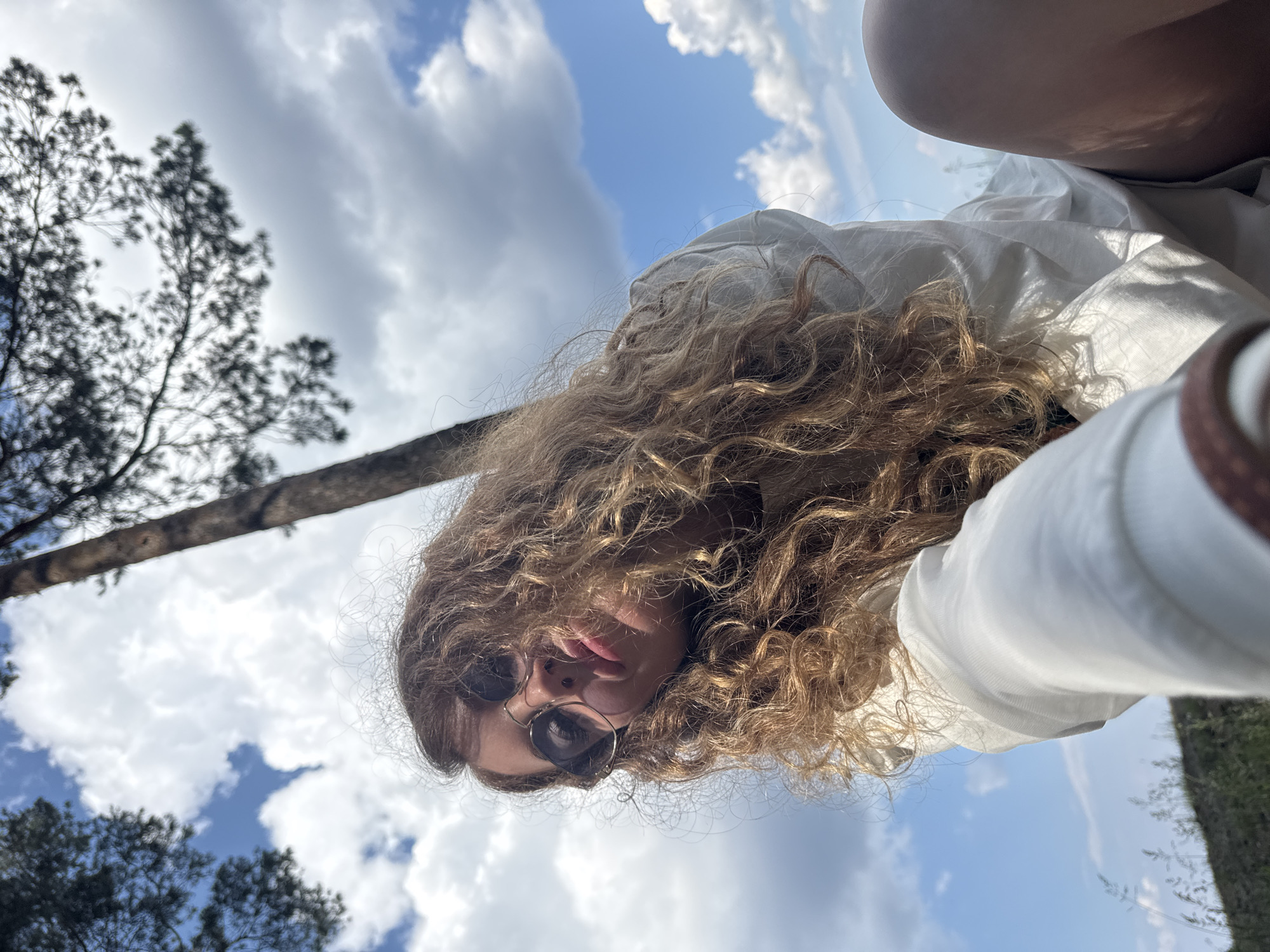 In this low-angle, upward shot, a young woman with long, curly blonde hair is captured outdoors, with a tree trunk and a bright blue sky filled with fluffy white clouds dominating the background. She is wearing round, dark-lensed sunglasses and a white garment. Her lips are pursed as if she is about to kiss or has just kissed. The environment appears to be natural, possibly a park or wooded area, given the presence of the tree and some vegetation visible at the bottom of the frame. The lighting suggests it is daytime, with sunlight illuminating the scene and casting shadows. The perspective implies the photo was taken by the woman herself, likely using a selfie mode, with the camera positioned low and pointing upwards. The image does not contain any identifiable landmarks or elements that would specifically place it within the city of Chaikovsky, Russia; the setting is generic to a natural outdoor environment.