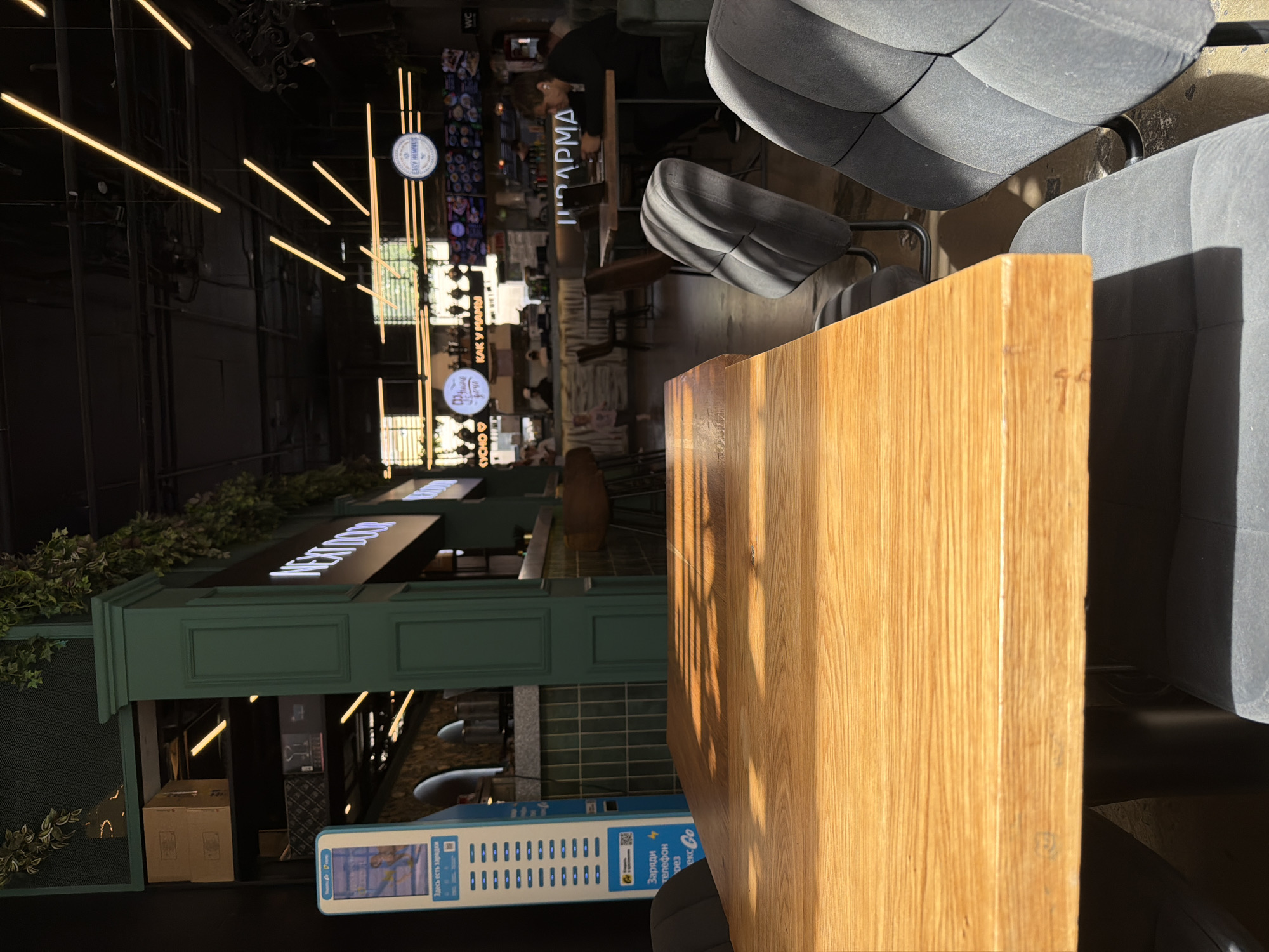 This is a photograph of the interior of a modern food court or restaurant, likely located in Saint Petersburg, Russia, given the Cyrillic text visible on signage and informational displays. The foreground is dominated by a large, polished wooden table with a visible wood grain pattern, catching shafts of sunlight that create strong shadows. To the right of the table, two upholstered grey chairs with curved backs are positioned, one partially obscuring the other.
In the middle ground, the scene opens up to a bustling food court environment. A green, pillar-like structure with decorative molding and artificial greenery on top is visible on the left. To its right, a charging station for mobile devices is prominently displayed, featuring Cyrillic text that includes "Заряди телефон" (Charge phone). Beyond this, there is a food counter with tiled green backsplashes and seating with wooden stools.
Further into the background, multiple food vendors are identifiable by their illuminated signage. One sign clearly reads "NEXT DOOR". Another sign, circular with text and an image, is visible above a food stall. A person, likely a customer, is seated at a table on the right, using a laptop and wearing earbuds. Another person, possibly staff, is behind the counter in the distance. The ceiling is a dark, industrial structure with numerous long, yellow fluorescent light fixtures diagonally arranged. The overall impression is of a bright, contemporary urban dining space.