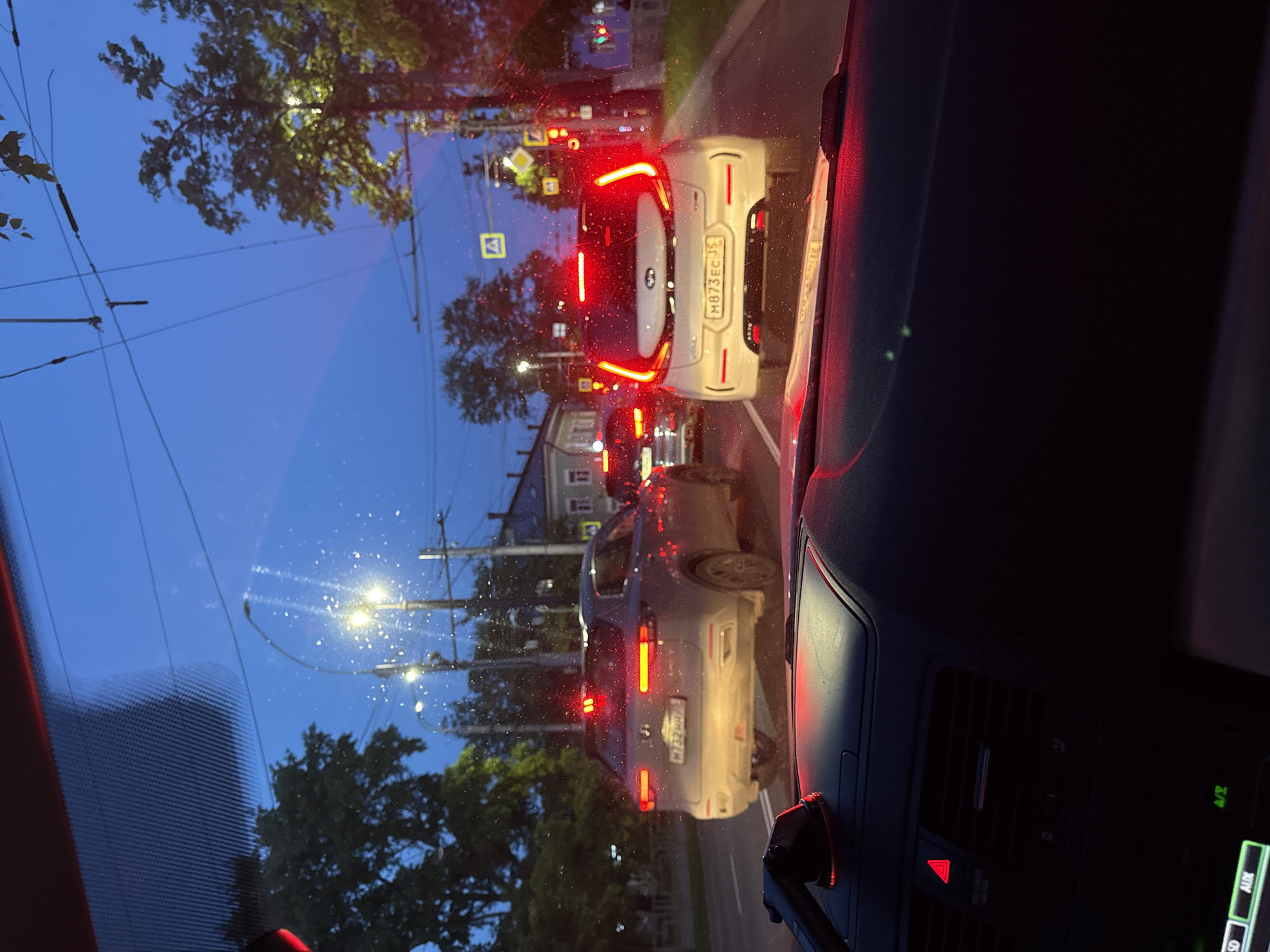 This image captures a street scene from inside a vehicle, looking through a windshield that appears to be wet with water droplets, suggesting recent rain. The time of day is dusk or early night, indicated by the dark blue sky, active streetlights, and illuminated car lights. The view is oriented vertically, with the sky and tree canopy on the left, and the street with cars on the right.
In the foreground, a white car is directly ahead, its bright red brake lights glowing. Its Russian license plate, `M873EC 35`, is clearly visible, with "35" indicating Vologda Oblast, Russia. Further along the road, another darker vehicle is visible, also with illuminated brake lights and a partially visible license plate beginning with `H212AA`. The cars appear to be in slow-moving traffic or stopped, possibly at an intersection, with traffic signs and a traffic light visible in the distance.
Overhead, a network of power lines or trolley lines stretches across the frame, partially obscured by tree foliage. Buildings with illuminated windows line the background. On the right and bottom edges of the image, elements of the car's interior are visible, including part of the dashboard with an air vent and a red triangle hazard light button. A screen displays "AUX" and other partial text like "PASSENGER AIR BAG" can be seen near the hazard button.