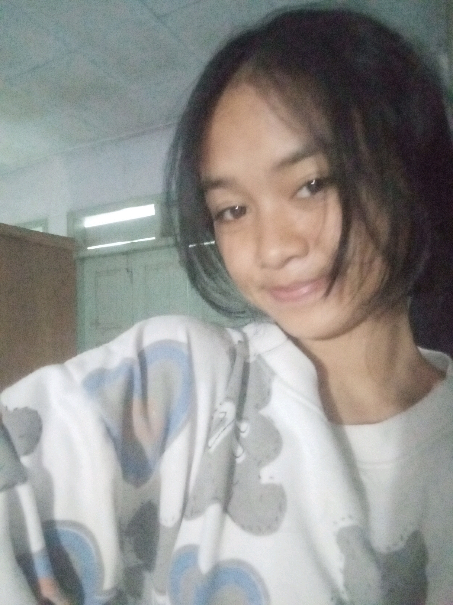 The image is a close-up, selfie-style shot of a young person with dark, short hair and dark eyes. They are looking towards the camera with a slight smile. The person is wearing a white sweatshirt with a gray and blue patterned design. The background appears to be an indoor setting, possibly a room, with a closet or cabinet visible on the left and a door or wall with light fixtures in the background. The lighting suggests it could be daytime or evening with artificial lights on. The scene conveys a casual and relaxed mood. There is no visible text in the image. The location context is Badas, Indonesia.