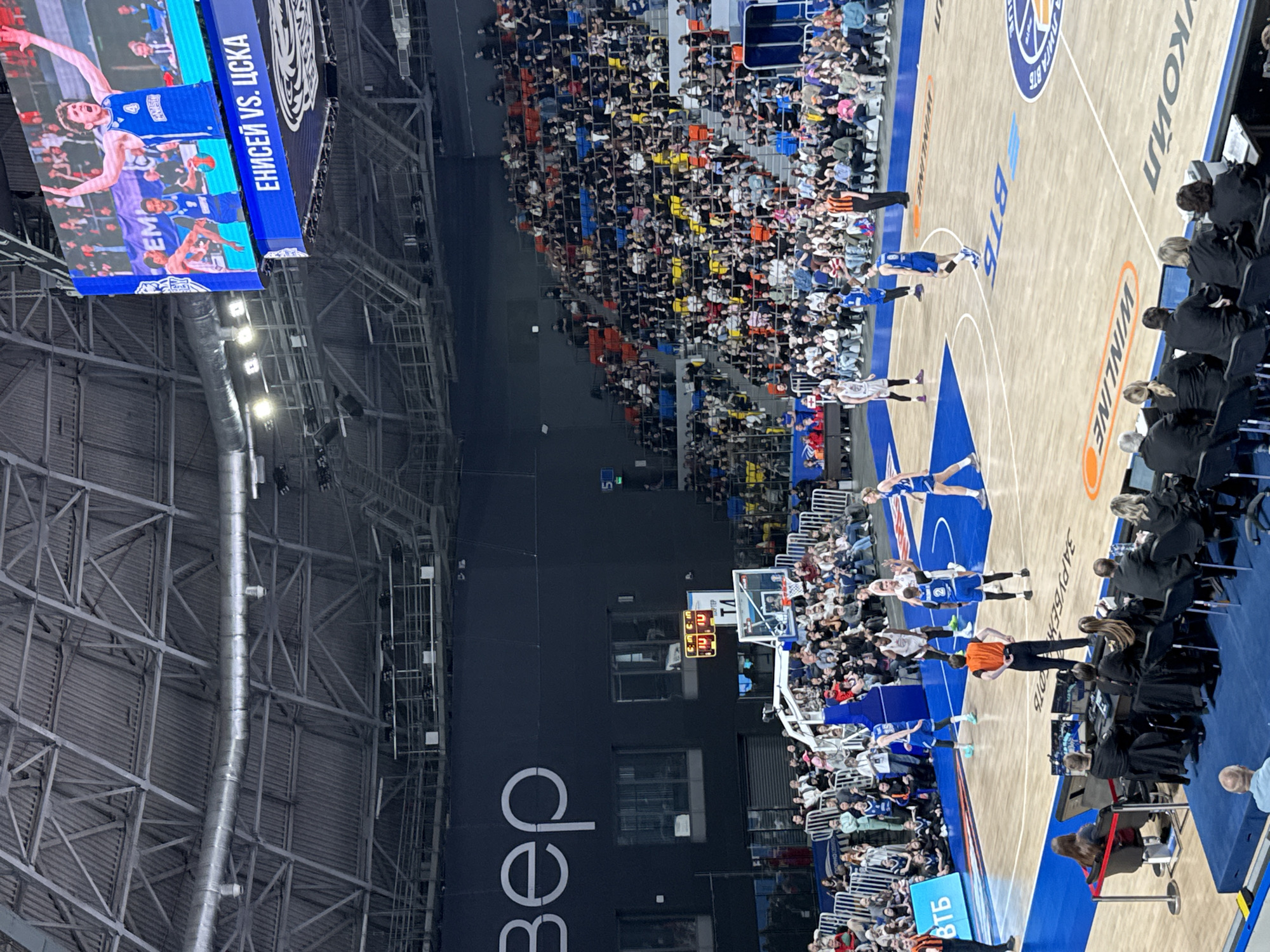 The image depicts a basketball game taking place indoors, with a large crowd in attendance. The location is likely in Krasnoyarsk, Russia, given the Cyrillic text on the scoreboard "ЕНИСЕЙ VS. ЦСКА" (Yenisey vs. CSKA) and the word "Вер" (Ver) which could be related to the city or a sponsor. The game appears to be in progress, with players on the court in white and blue uniforms. There is an official in an orange shirt positioned near the players. The court has advertising markings, including the word "WINLINE" and "BTB". The scoreboard shows numbers related to the game score or time. Spectators fill the stands. The arena features visible metal ceiling supports.