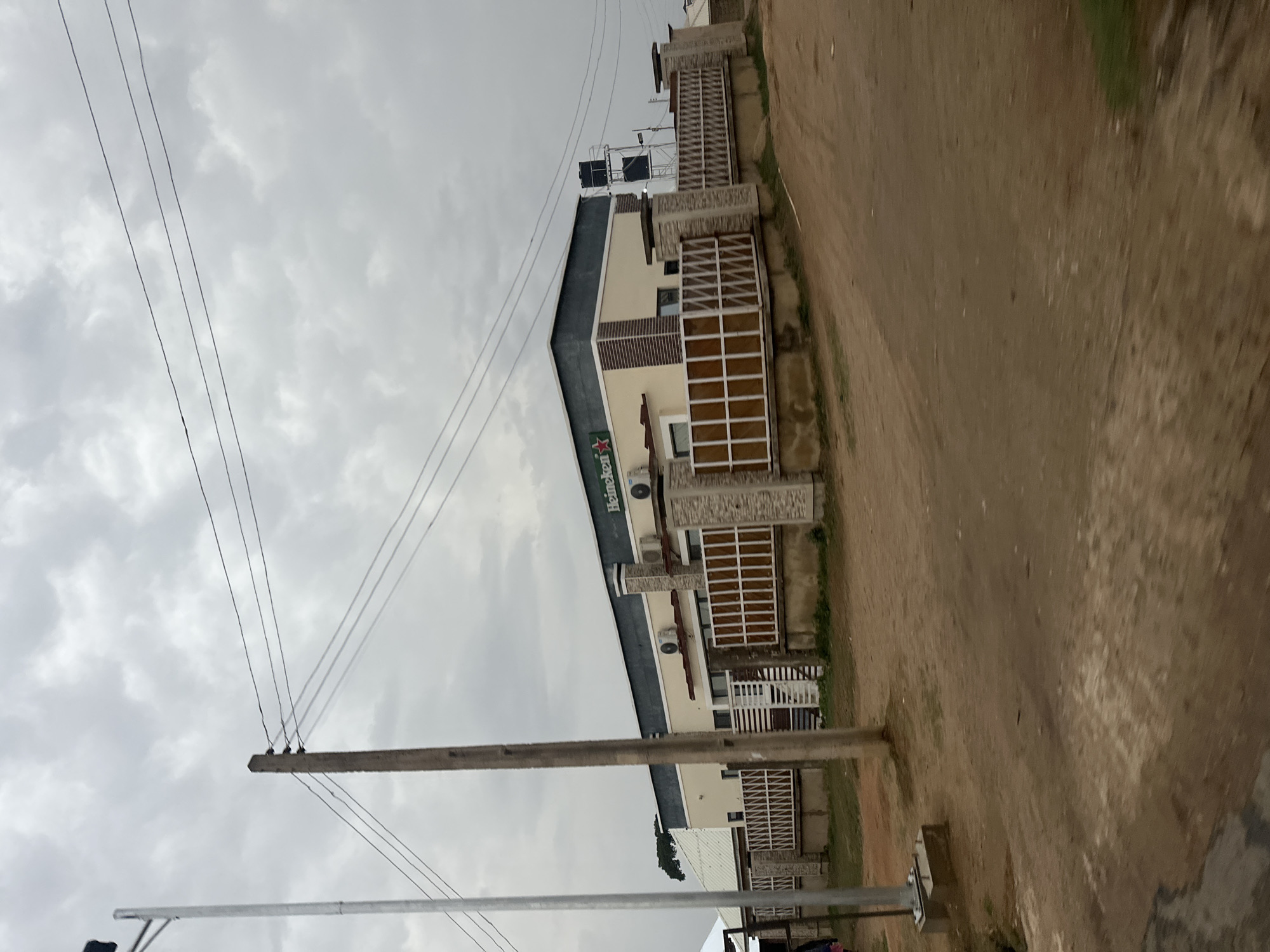 The image depicts a building with a Heineken sign in Jos, Nigeria. The building has a beige facade with brown accents and a dark gray roof. It is a multi-story structure with decorative window coverings. Utility poles with electrical wires are visible in the foreground, set against a cloudy sky. The ground is dirt or gravel. The scene appears to be taken from a roadside perspective.