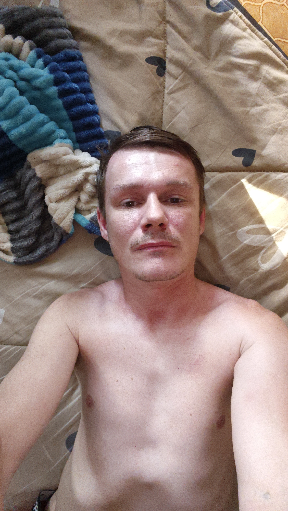 A bare-chested man lies on a patterned blanket with dark hearts. A blanket with blue, grey, and white stripes is to his left. The man has light skin, brown hair, and a light mustache and goatee. The scene appears to be indoors, possibly a bedroom. The location is Kaltan, Russia.