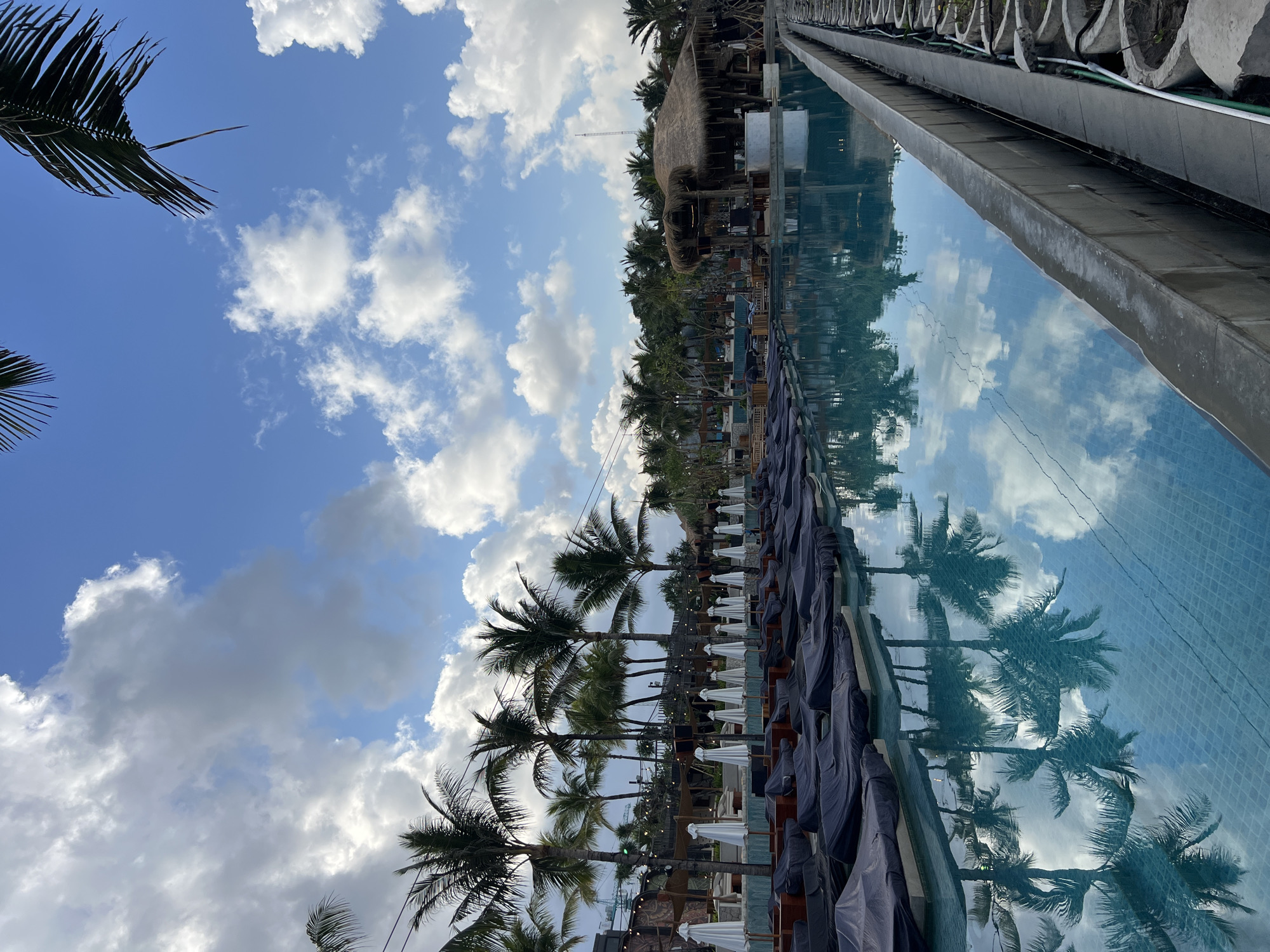 The image shows a swimming pool at a resort in North Kuta, Indonesia. The pool is empty and reflects the sky, clouds, and palm trees. There are lounge chairs covered with dark fabric along the edge of the pool. In the background, there are more palm trees, a thatched-roof bar, and some buildings. The sky is blue with white clouds.