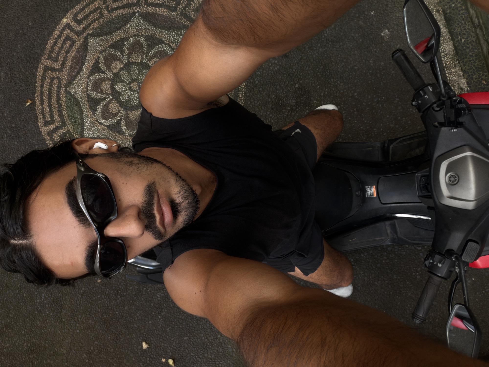The image is a high-angle selfie taken in North Kuta, Indonesia. A man with brown skin, black hair, a beard, and sunglasses is sitting on a motorcycle. He is wearing a black t-shirt and dark shorts. White earbuds are visible in his left ear. The motorcycle is black and gray, with red accents on the seat. The ground is paved with gray stones, and a circular pattern is visible on the left side of the frame. The man is looking directly at the camera, holding the device to capture the image.