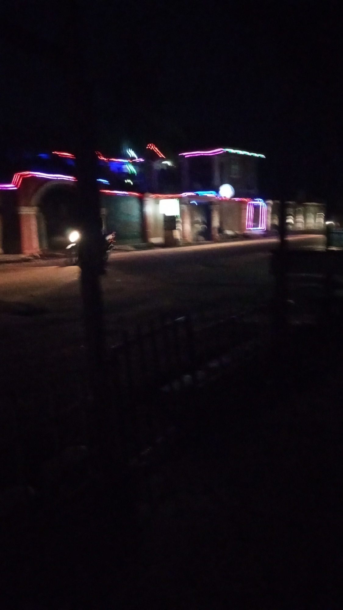 This nighttime image, set in Narayi, Nigeria, captures a street scene illuminated by colorful decorative lights. The upper portion of the frame is dominated by darkness, indicative of night. Buildings line what appears to be a street or pathway, adorned with vibrant string lights in shades of red, blue, pink, and green, outlining their structures and facades.
On the left, a building features an arched entryway highlighted by these lights. Moving rightward along the street, other structures are similarly decorated, with one displaying a circular glowing object and another featuring a prominent, bright rectangular frame made of lights, possibly marking an entrance or window.
In the midground, a paved road stretches across the scene. A vehicle, likely a motorcycle or scooter, is visible on the left side of the road, identified by its glowing headlight. The image exhibits significant motion blur, especially noticeable in the streaked appearance of the lights and the vehicle, suggesting either camera movement during capture or a moving subject in low light. A dark, low fence or barrier is partially visible in the immediate foreground. The absence of natural light, combined with the artificial illumination, defines the nocturnal setting. There are no visible people or discernible text within the frame.