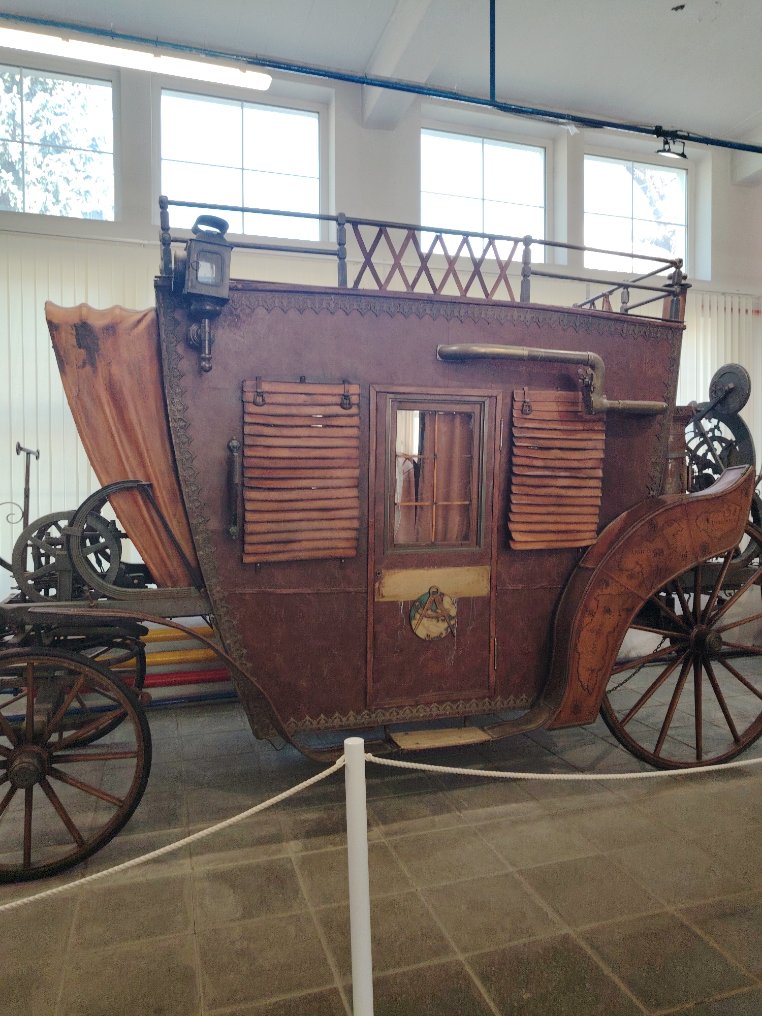 This media file captures a detailed view of a large, antique-style carriage or stagecoach, displayed indoors, likely in a museum or exhibition hall in Moscow, Russia.
The carriage itself is the central subject, primarily dark brown in color, possibly made of aged leather, wood, or a similar material, and features intricate decorative detailing along its edges. It rests on large wooden spoked wheels, two of which are clearly visible. A faded, light brown fabric or canvas canopy is draped towards the rear-left.
The side of the carriage boasts a window with an interior curtain and a section covered by horizontal slatted blinds or shutters. A small, dark lantern is mounted near the front top of the carriage, below a metal railing with a crisscross pattern along the roofline. A brass-colored pipe-like structure is also visible on the upper side. Below the window, there's a circular emblem featuring what appears to be a compass and other navigational instruments. Further back, on a curved wooden panel near the rear wheel, a faded map-like design is etched, with partially legible words like "MEXICO" and "ABIA."
The setting is bright and well-lit, with natural light streaming through several large rectangular windows set high in a white wall. Overhead, fluorescent lights and blue pipes run along the ceiling. The floor is composed of large, grey square tiles. A simple white rope barrier, supported by white posts, separates the carriage from the viewer, reinforcing the impression of an exhibit. In the background, behind the carriage, colorful horizontal bars (red, yellow, blue) are faintly visible.
No people are present in the image, and the carriage is stationary, serving as a historical display. The time of day appears to be daytime due to the natural light.