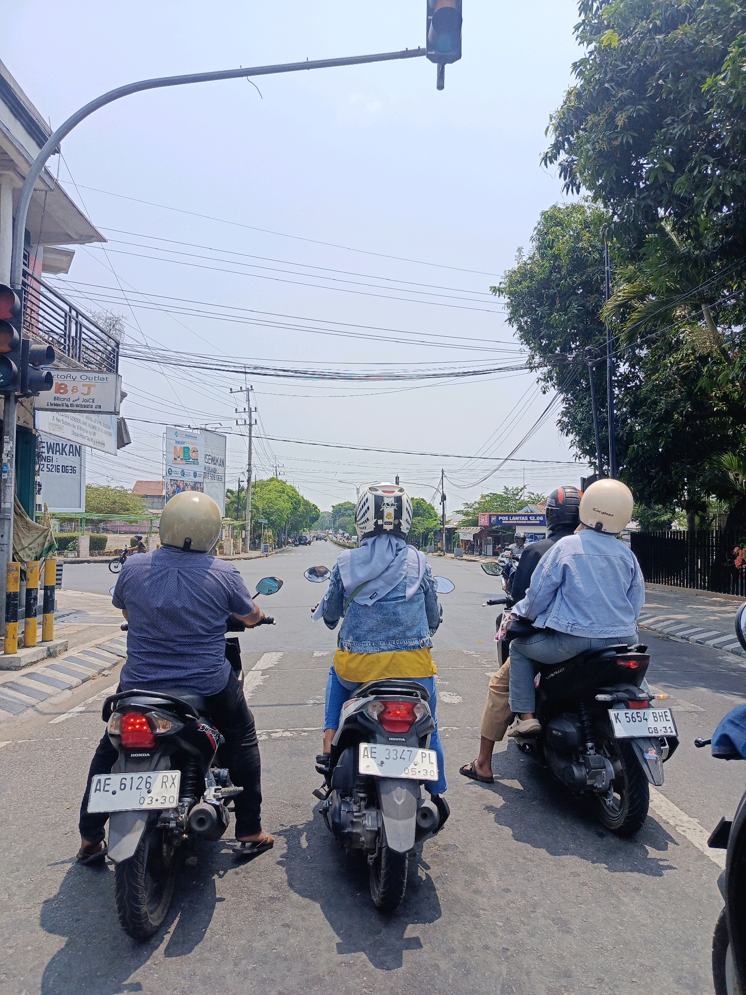 The image depicts a street scene in Sukowinangun, Indonesia, on a bright, clear day. The perspective is from behind three individuals riding motorcycles in a line, facing forward towards a road intersection. The foreground features the backs of the motorcycles and riders. The rider on the left, wearing a blue patterned shirt and a cream-colored helmet, is on a black Honda motorcycle with the license plate AE 6126 RX. The middle rider, in a denim jacket and a white helmet with black accents, is on a light-colored motorcycle. The rider on the right, wearing a light blue denim jacket and a cream helmet, is on a black Vario motorcycle with the license plate K 5654 BHE. Both middle and right riders appear to be a pair.
The environment includes a traffic light suspended over the intersection, a building on the left with a "Factory Outlet" sign, and several billboards and power lines overhead. Trees line the sides of the road, and other vehicles are visible further down the road, suggesting moderate traffic. The road surface is asphalt with painted lane markings. The overall impression is a typical urban street scene in Indonesia during daylight.