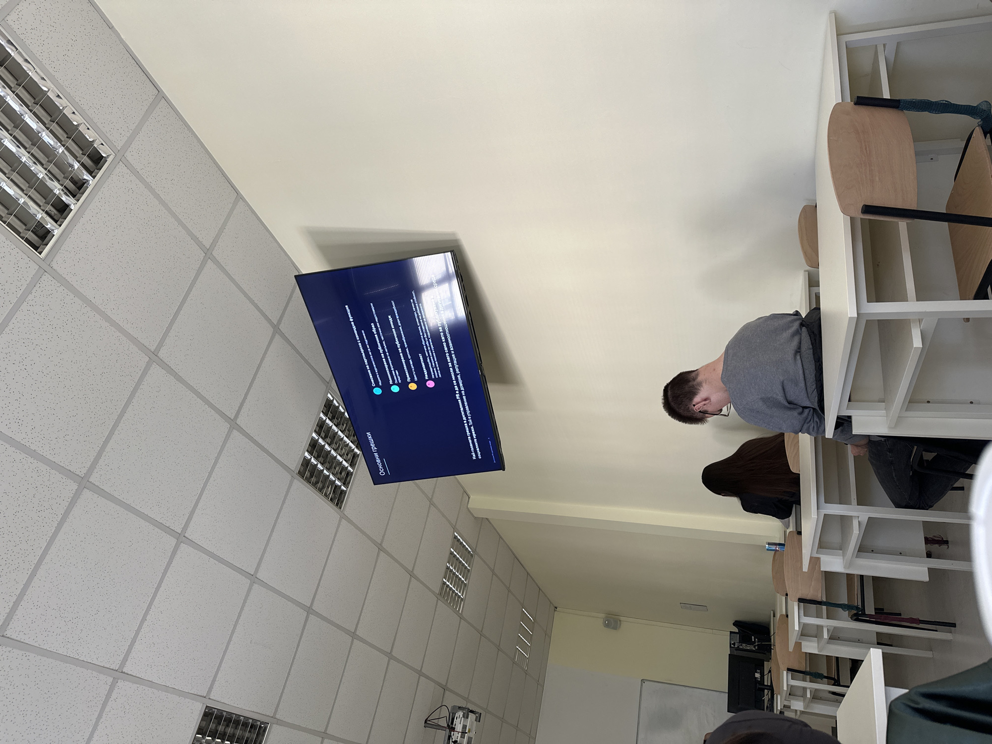 The image shows a classroom setting with students sitting at desks and a TV screen displaying text in what appears to be Bulgarian. The text includes the phrases "Основни грешки" and "Най-голямата грешка в дигиталния PR". The classroom is located in a building that has a suspended ceiling with recessed lighting. The location is likely in Sofia, Bulgaria, given the text displayed.