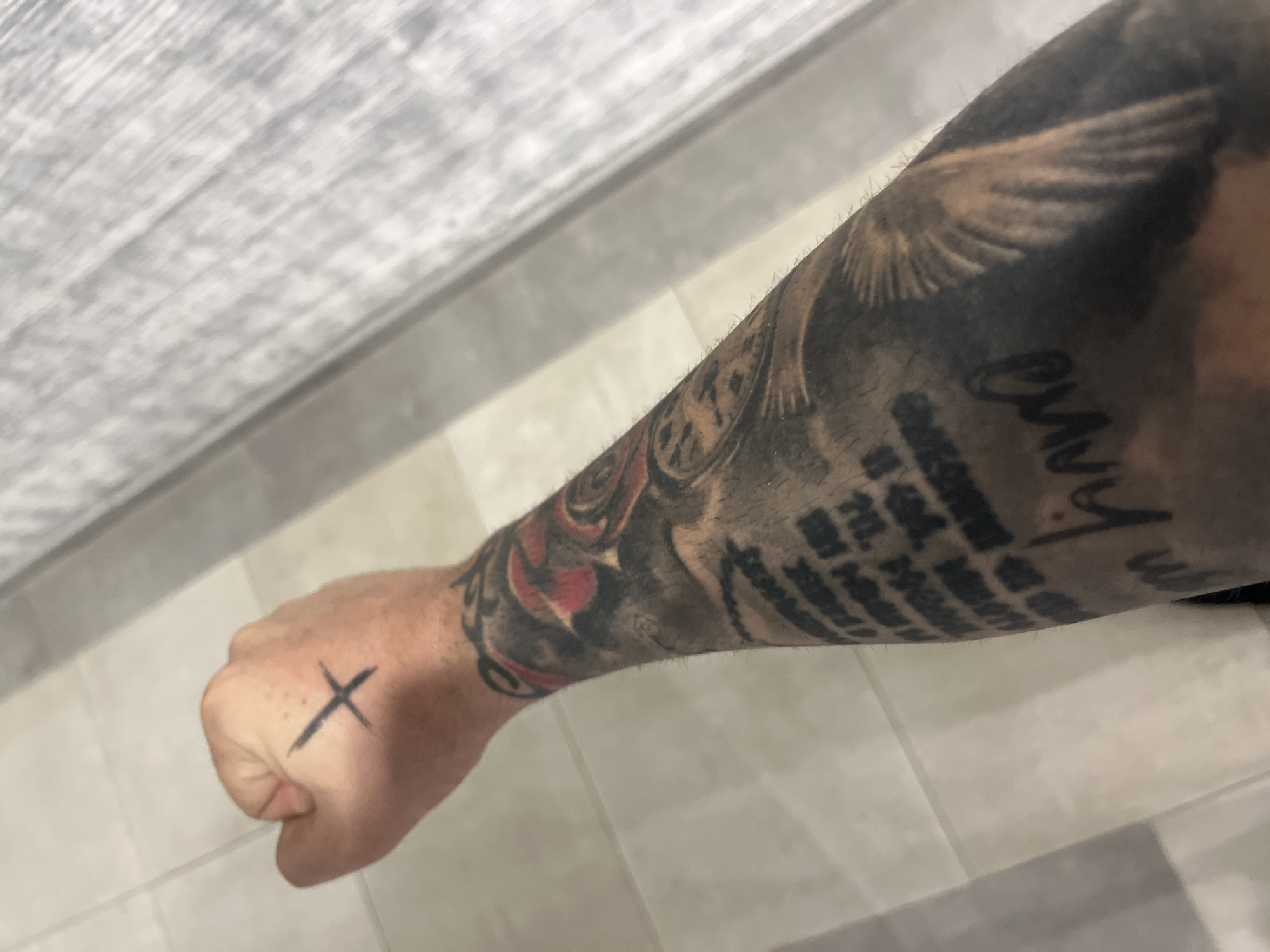 A tattooed forearm and hand are visible, resting on a tiled floor. The hand, clenched into a fist, has a black cross tattoo on the back. The forearm is heavily tattooed with what appear to be text, a clock face, and a wing. The background consists of light-colored square tiles and a blurred, grey, slatted surface. The image does not provide any information to indicate its location in Varna, Bulgaria.