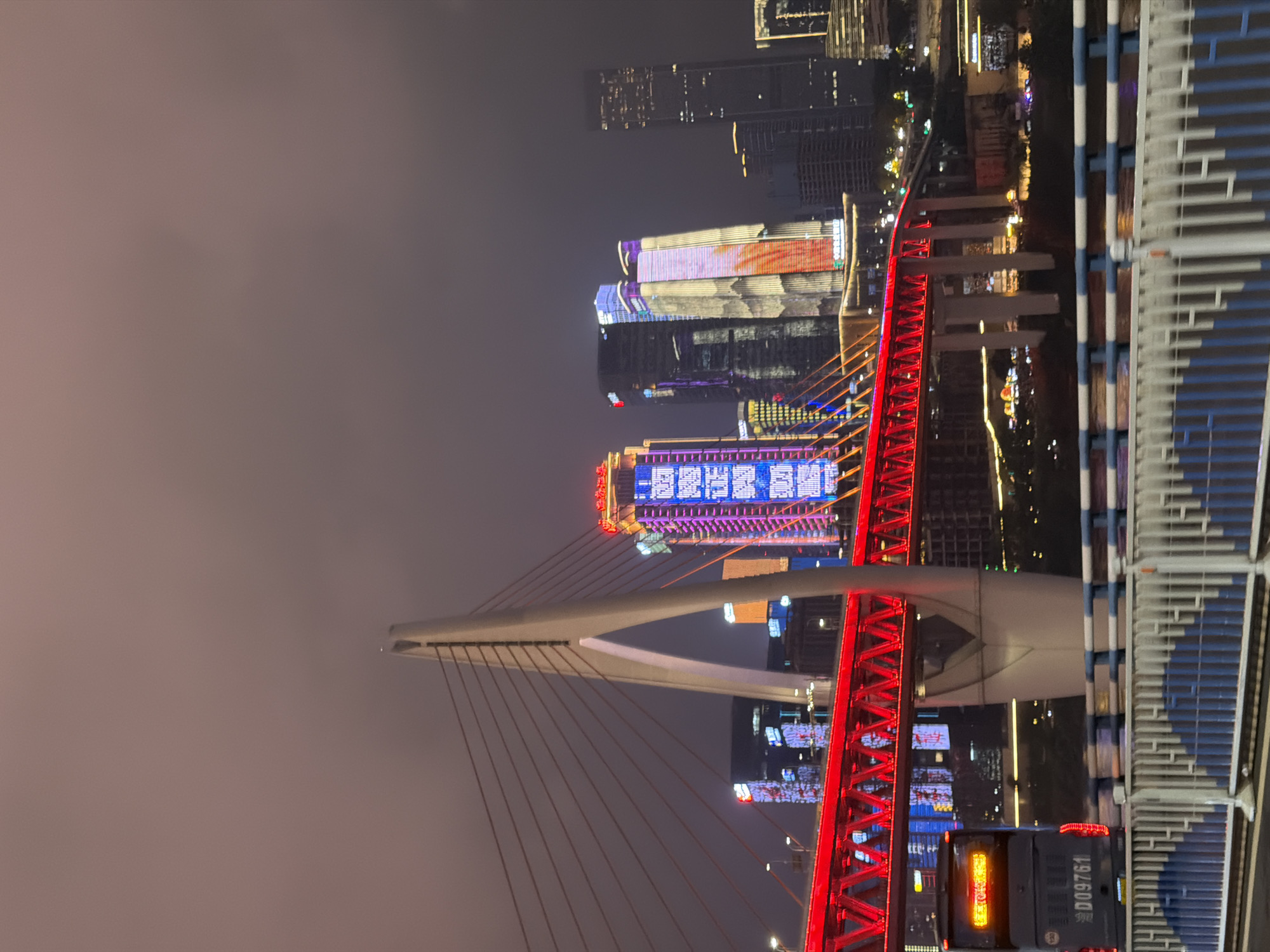 This image depicts a nighttime view of a modern cityscape in Yuzhong District, China, characterized by a prominent cable-stayed bridge and illuminated skyscrapers. The bridge features a stark white pylon supporting numerous red cables that extend towards the towers of the buildings. The bridge deck is also illuminated in a vibrant red. Several tall buildings are visible, adorned with colorful LED lights, creating intricate patterns and displaying text. One building in the foreground displays a large vertical LED screen with purple and white text, while another adjacent building features a similar screen with a colorful, abstract design of orange and white. A bus with illuminated signage is partially visible on the bridge, suggesting traffic flow. The sky is overcast and dark, indicating a cloudy evening or night. The foreground includes a section of a white and blue metal railing, suggesting the photograph was taken from a vantage point on another bridge or elevated walkway. The water body below the bridge is visible, reflecting some of the city lights.