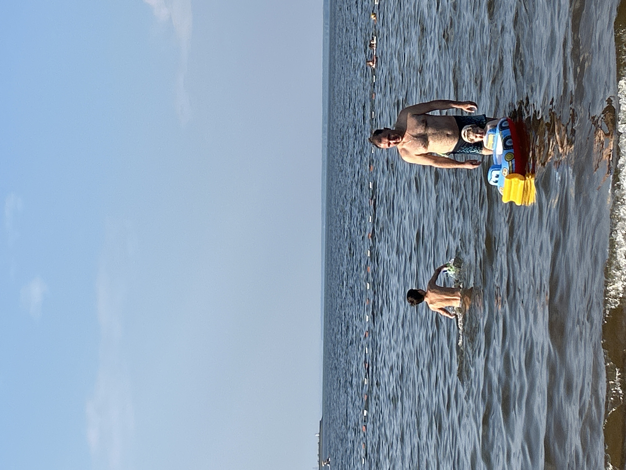 In Chaikovsky, Russia, on a bright, clear day, a man and two children are in the water. The man, wearing swim trunks, stands waist-deep in the dark blue water. To his right, a child sits in a blue and yellow toy boat. Further to the left, a second child, shirtless, is also in the water. A rope with buoys stretches across the water in the background. The horizon is a hazy line where the water meets the sky, which is a pale blue with a few wispy clouds.
