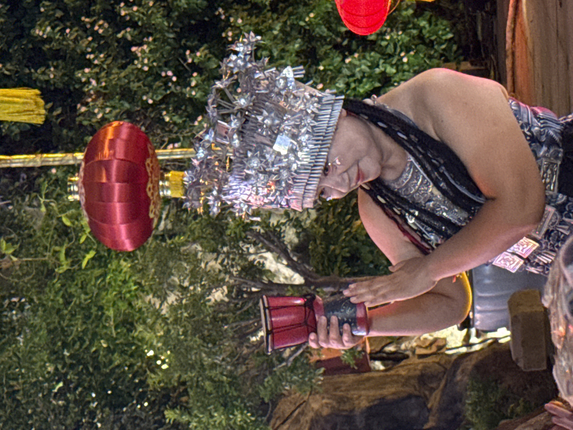 In Yuzhong District, China, a woman adorned in a silver headdress and traditional attire is depicted holding a small red drum. She stands against a backdrop of lush green foliage and a vibrant red lantern. The woman is captured mid-motion, with her hands near the drum, suggesting a performance or dance. The scene is illuminated, with warm light casting shadows and highlighting the intricate details of her costume and the surrounding environment.