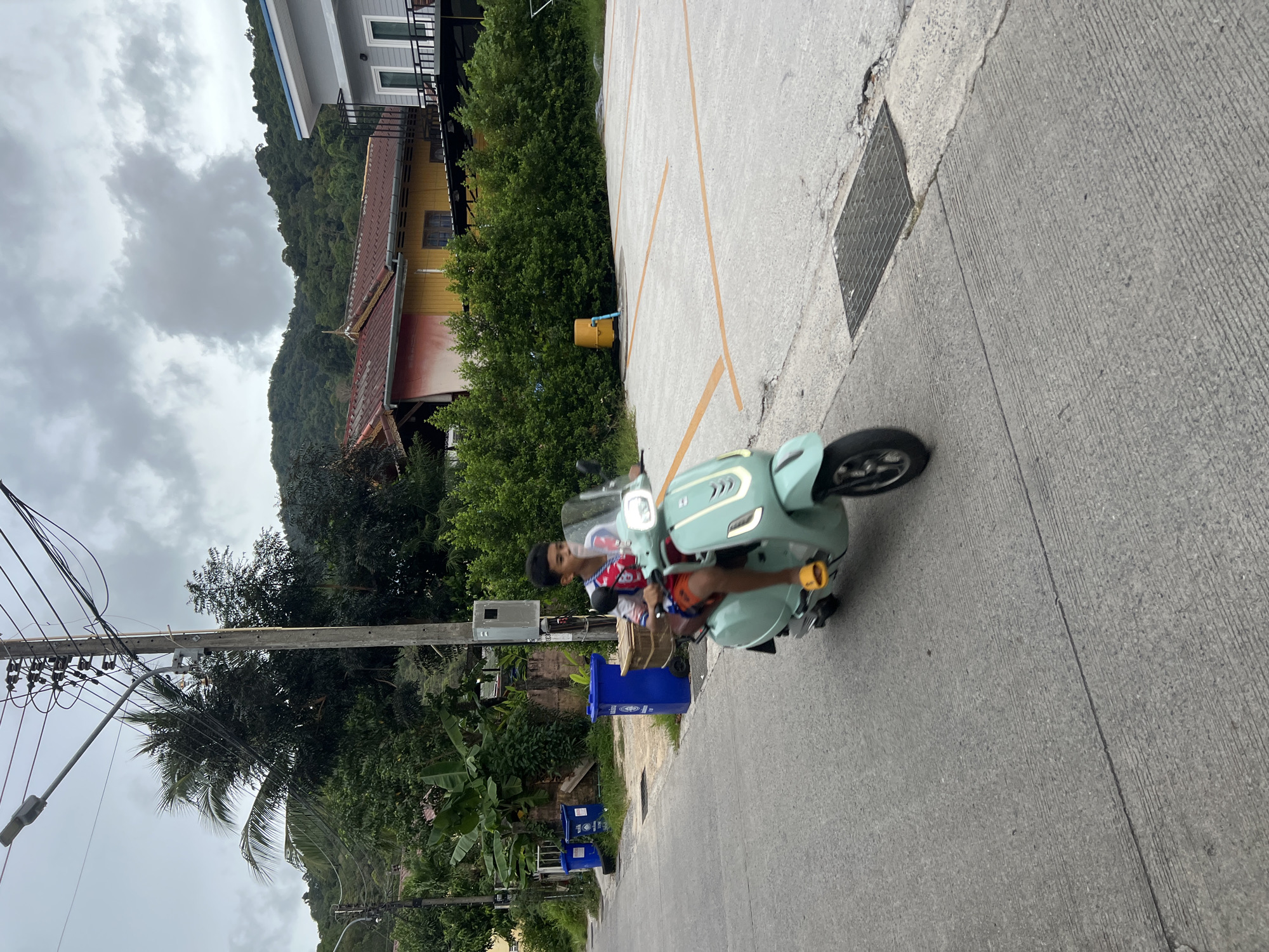 A young boy is riding a light teal scooter down a paved street in Choeng Thale, Thailand. He is wearing orange shorts, a red and white shirt, and yellow sandals. The boy is looking forward as he rides. The street is lined with trees and bushes on the left side. There are several blue trash cans on the left side of the street. In the background, there are houses and a hill covered in trees. The sky is cloudy.